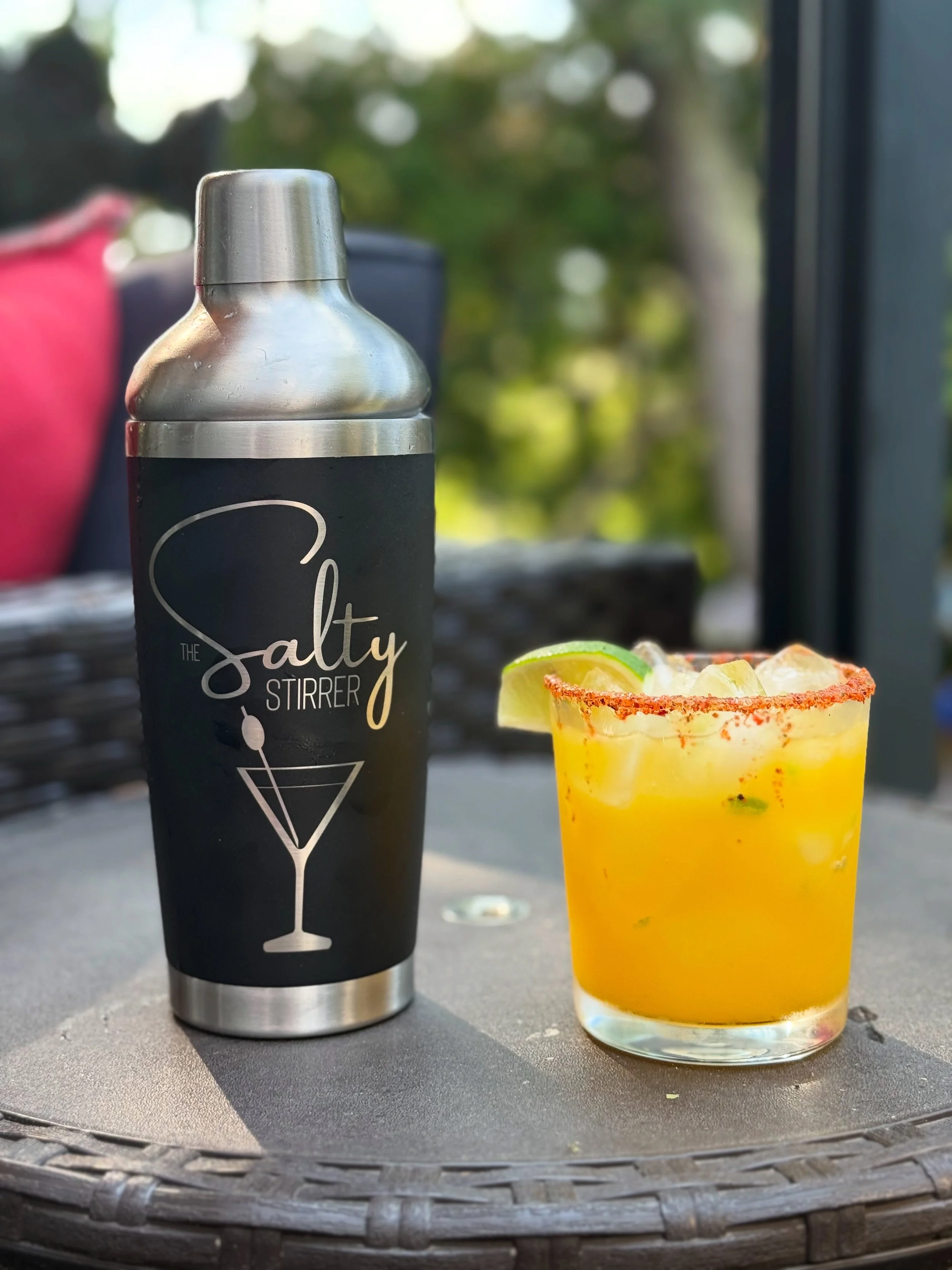 A stainless steel cocktail shaker with a black label reading 'Salty Styler,' placed on an outdoor table next to a glass of yellow cocktail garnished with lime and chili salt rim, with blurred greenery in the background.