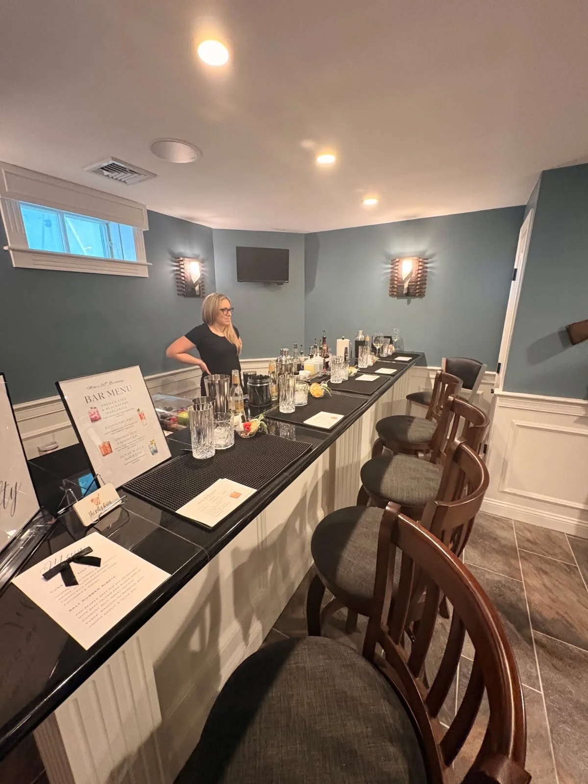 Bar area with a woman standing behind the counter, bar menu, glasses, and bottles on the countertop, chairs in front, interior with blue walls, wall-mounted lights, window, and TV.