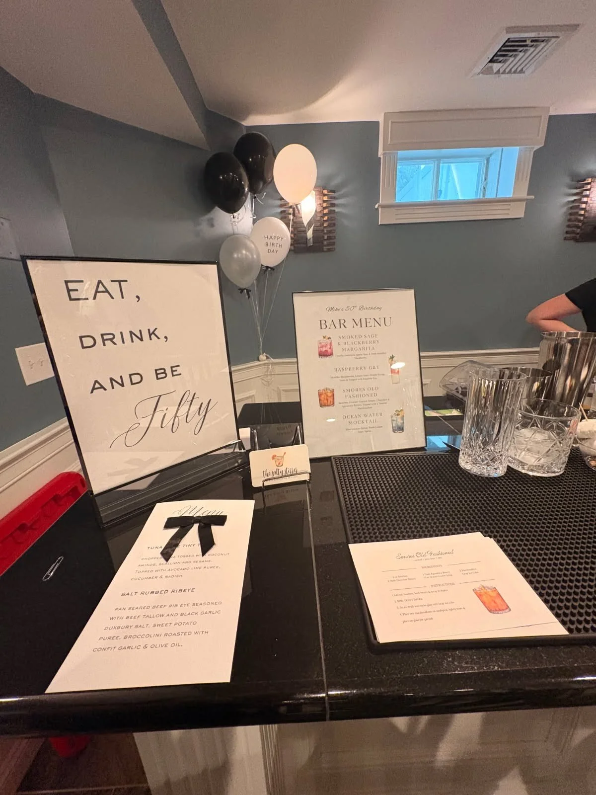 Signs and menus on a bar counter at a birthday celebration, with black and white balloons and a mural on the wall in the background.