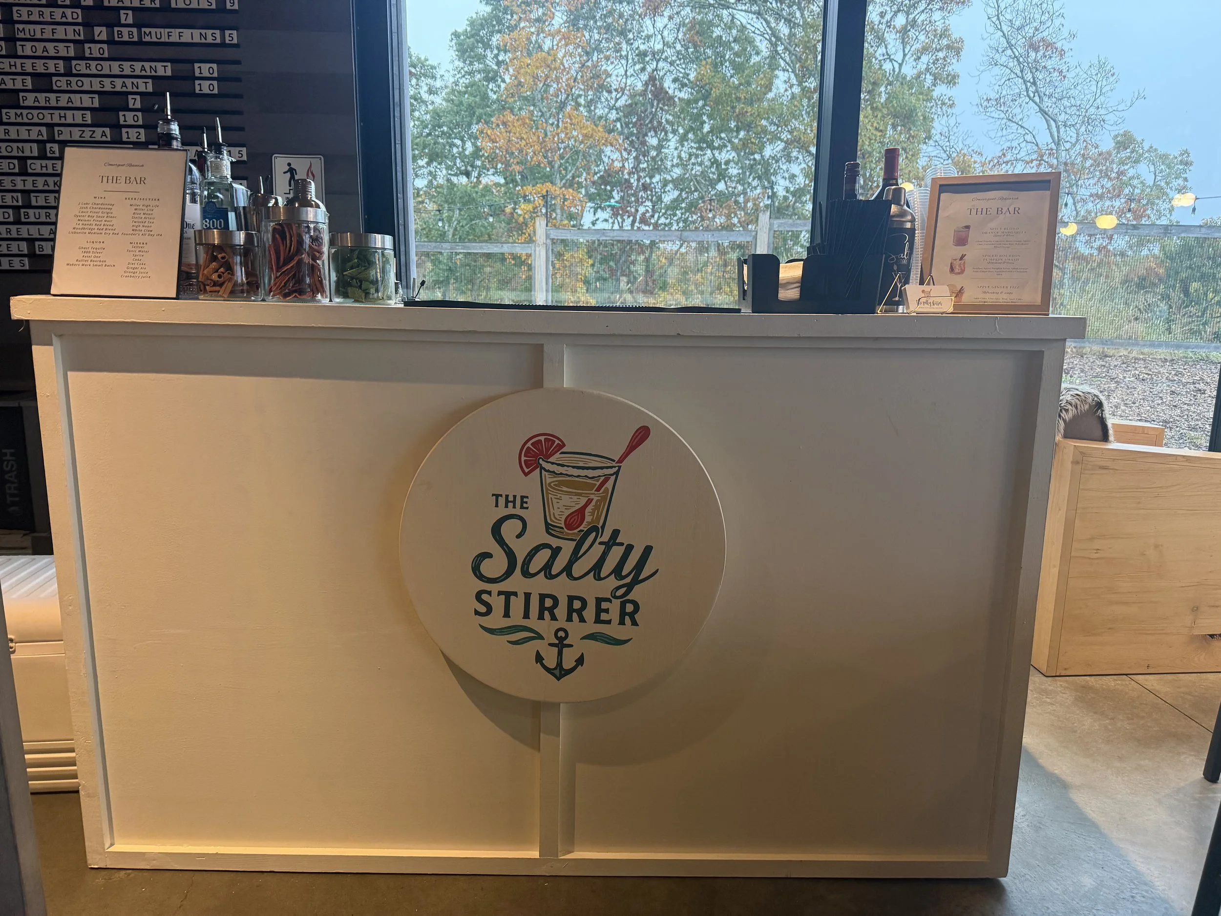A counter at a smoothie bar with jars of various ingredients, a menu board, and promotional frames. The counter features a sign that reads 'The Salty Stirr' with an anchor logo, and a large window behind shows trees with fall foliage.