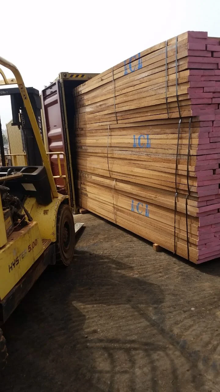 A forklift is carrying large stacks of lumber with pink edges, tied together with black straps, on a dirt surface.