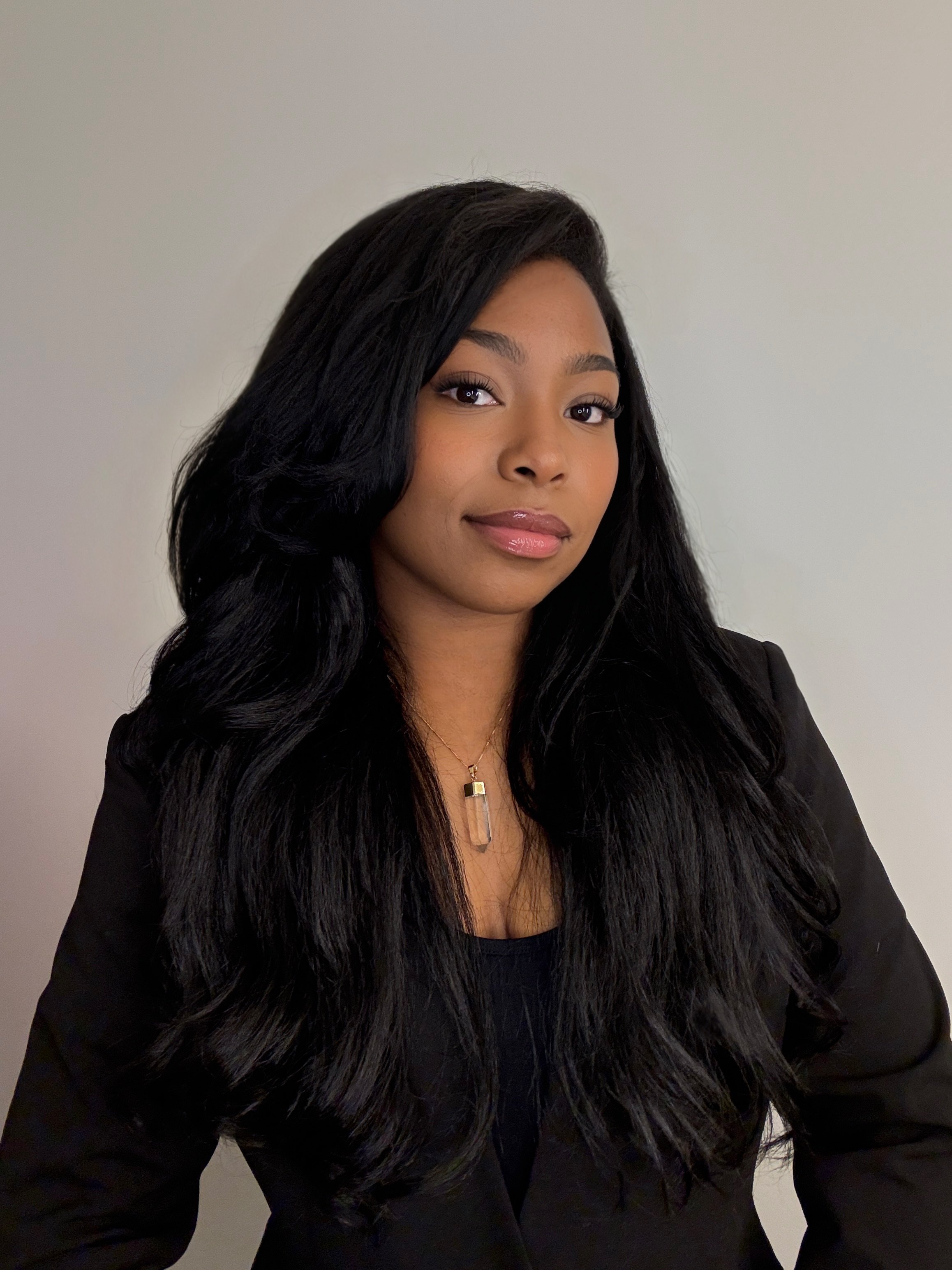 A woman with long black hair, wearing a black blazer and a necklace with a clear pendant, standing against a plain light-colored wall.