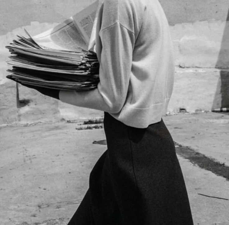 Person walking outdoors carrying a large stack of newspapers or magazines.