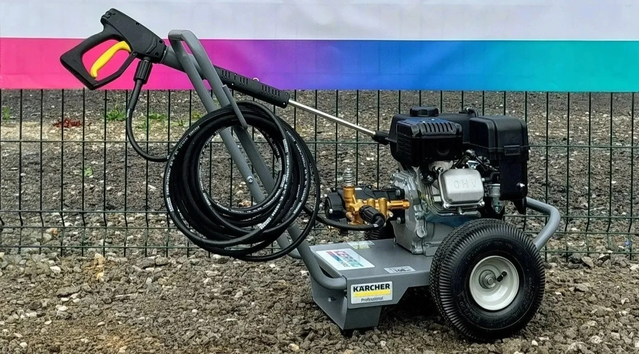 A high-pressure washer with a black engine, a yellow trigger gun, and hose, mounted on grey wheels, placed outdoors on gravel, with a fence and a rainbow-colored banner in the background.