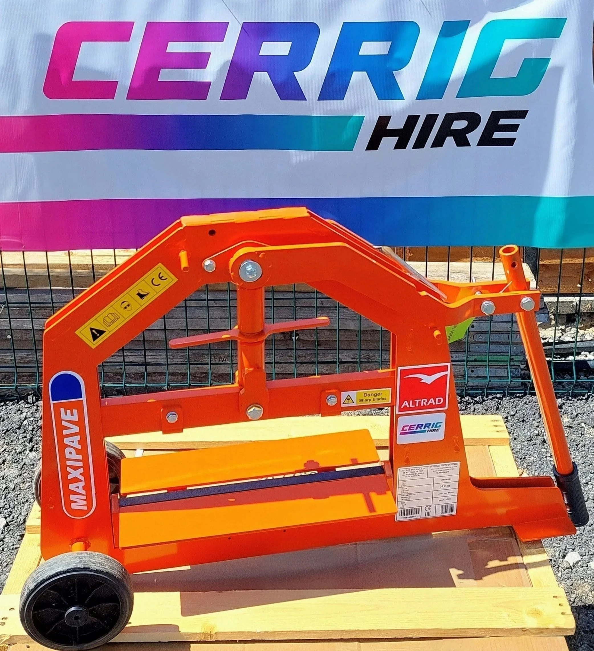 Bright orange block splitter with warning labels, positioned on a wooden surface outdoors. Behind it is a colorful banner that reads 'CERRIG HIRE' in bold purple, blue, and black letters.