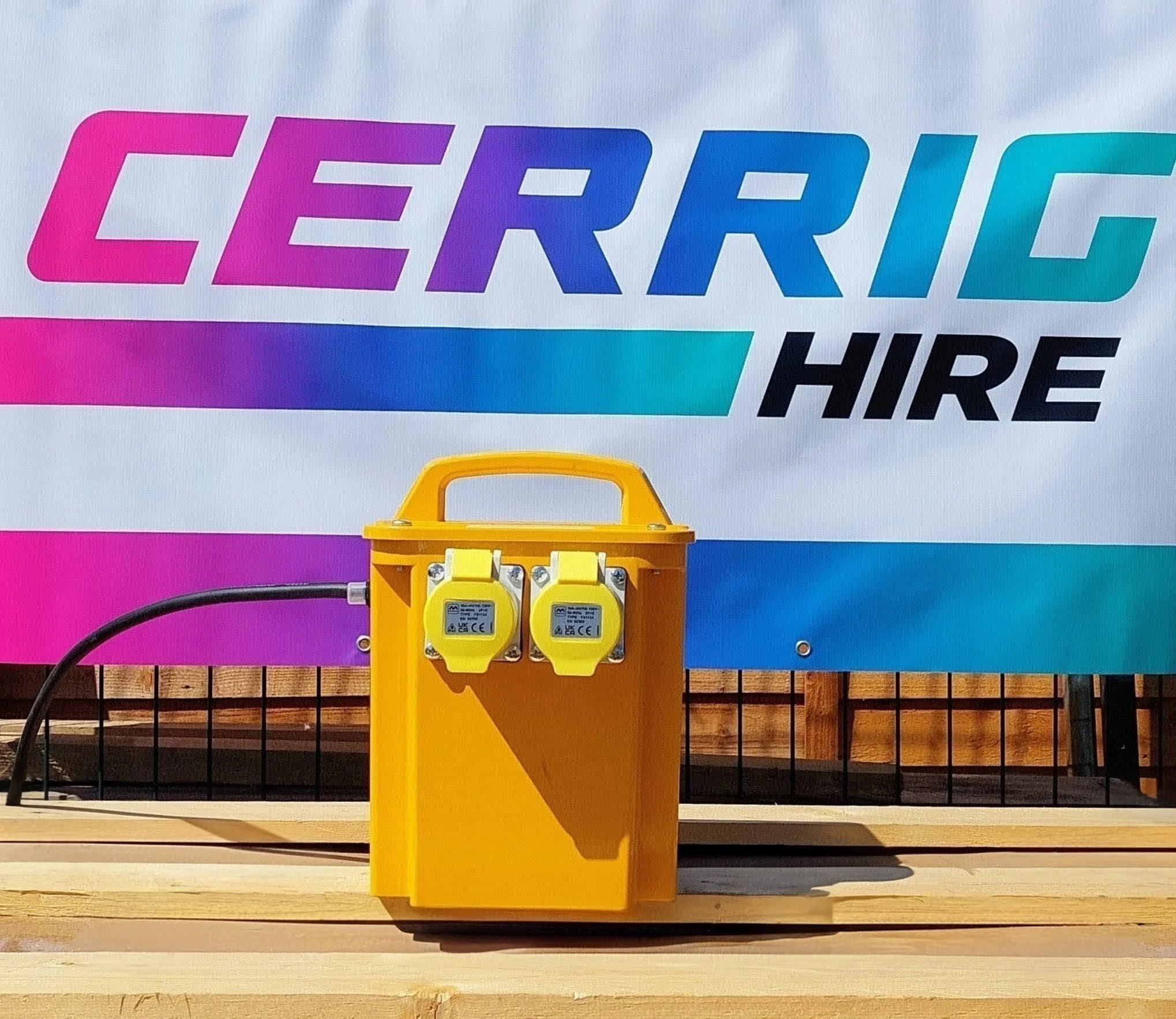 Colorful sign with the words 'CERIG HIRE' in bold letters, attached to a yellow electrical outlet box with cords plugged in, mounted on a wooden surface.