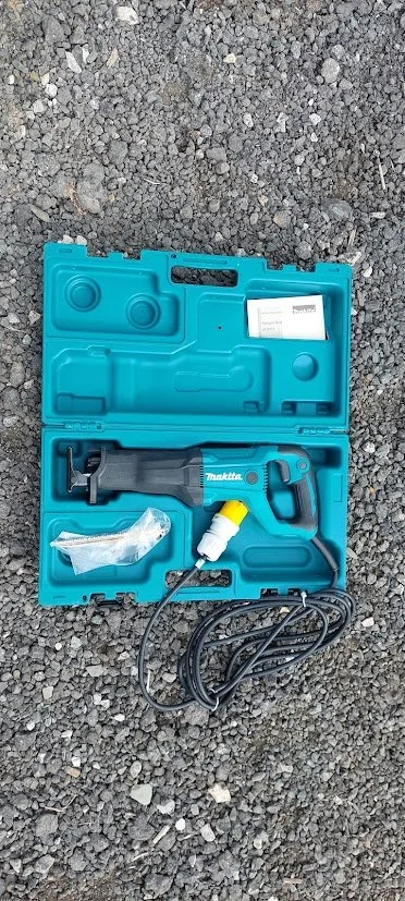 A power tool kit inside a blue plastic case, placed on gravel ground, containing a cordless reciprocating saw, a yellow and white accessory, and a power cord.