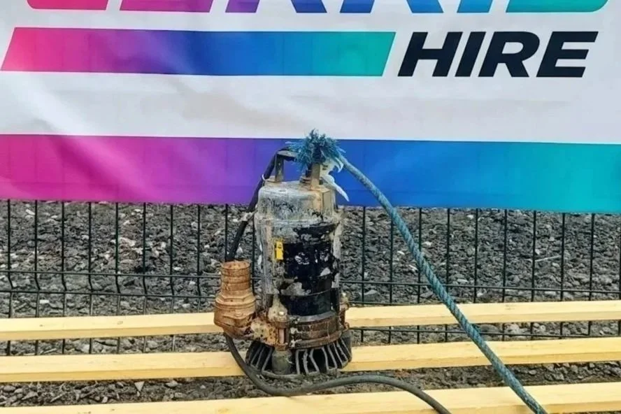 A submersible pump with a blue rope attached, placed on wooden planks in front of a fence and a colorful banner.