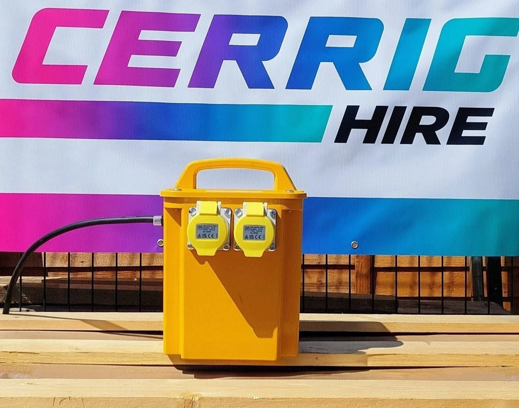 An electrical panel with black cable, mounted on a yellow support, located in front of a colorful wall with text that says 'CERRIG HIRE' in gradient shades of pink, purple, blue, and cyan.