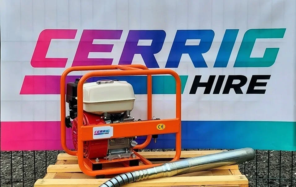 Portable petrol-powered concrete vibrator with orange frame, placed on wooden surface outdoors, with a large banner in the background reading "CERRIG HIRE".