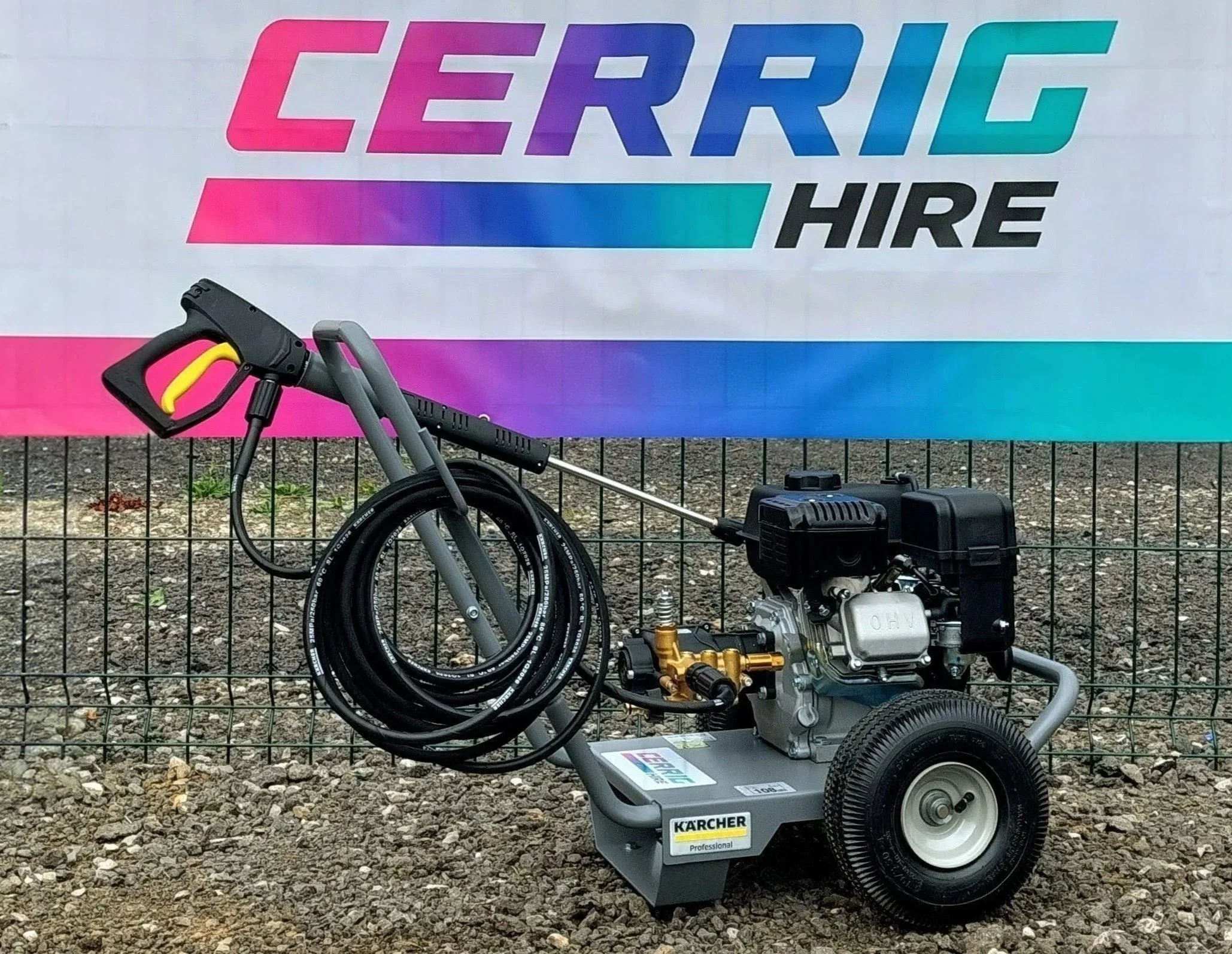A high-pressure washer machine with hoses and a power unit, positioned in front of a colorful sign that reads 'Cerrig HIRE' with a gradient background of pink, purple, and teal, and a black metal fence in the foreground.