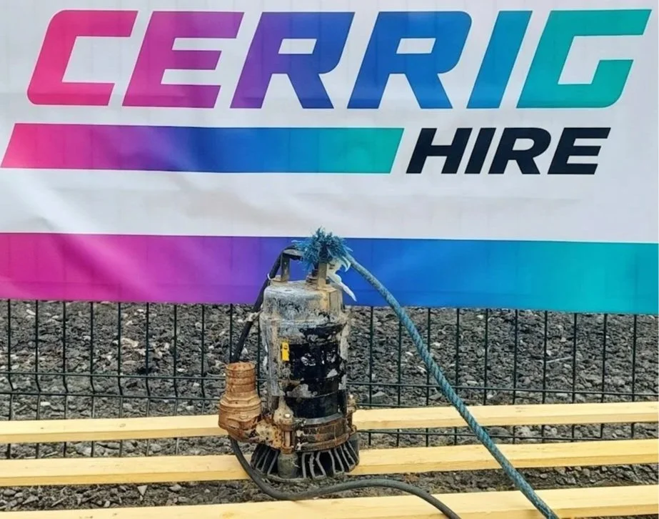 A water pump connected to a blue and black power cord on a wooden pallet, in front of a sign that reads 'CERRIG HIRE' in colorful letters.