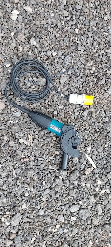 An electric angle grinder with a long power cord and a white and yellow electrical connector, lying on gravel ground.