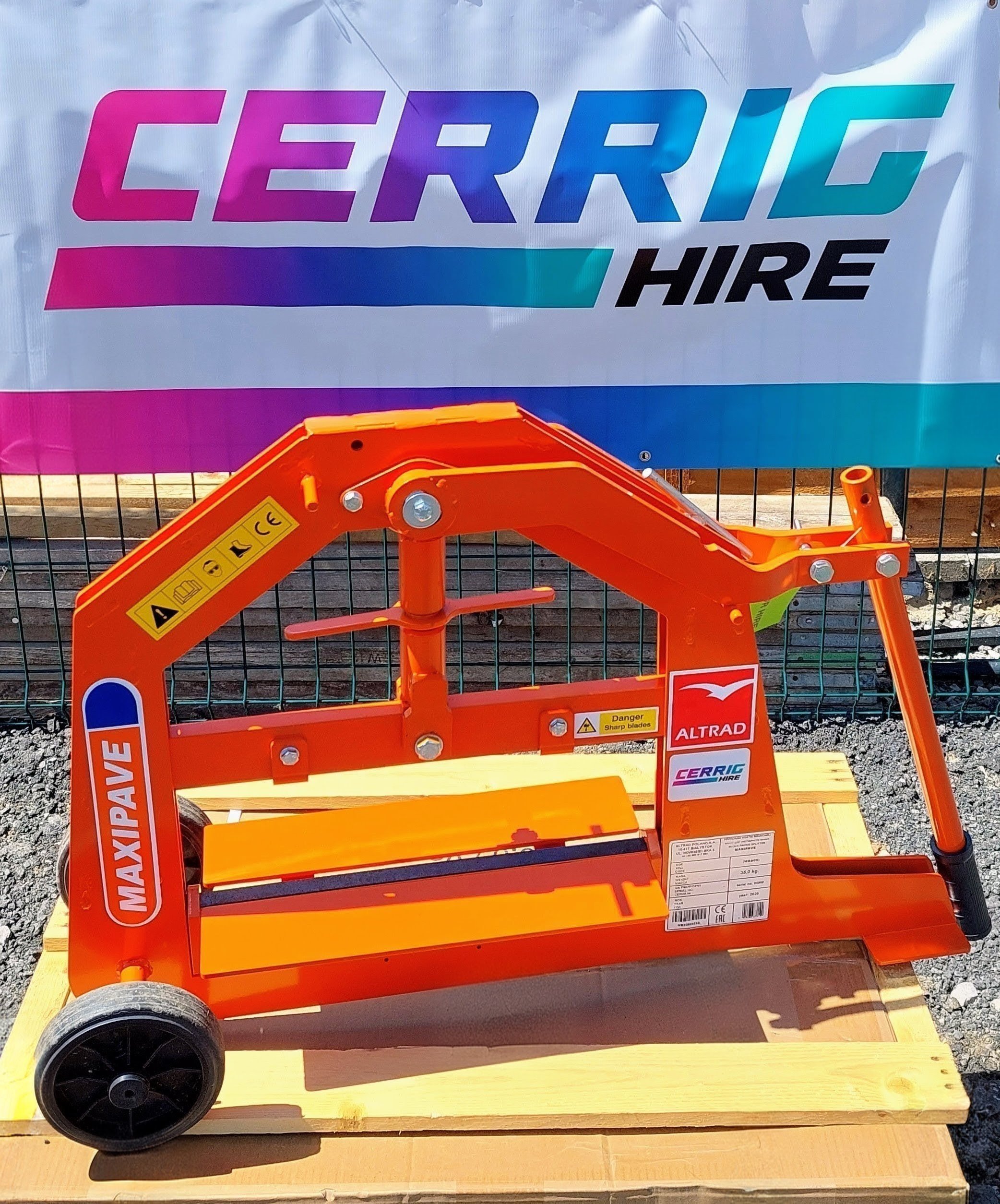 A bright orange block splitter with black wheels is placed on a wooden platform. A large white banner with colorful text is in the background, attached to a wire fence. Some small gravel is visible on the ground in front of the platform.