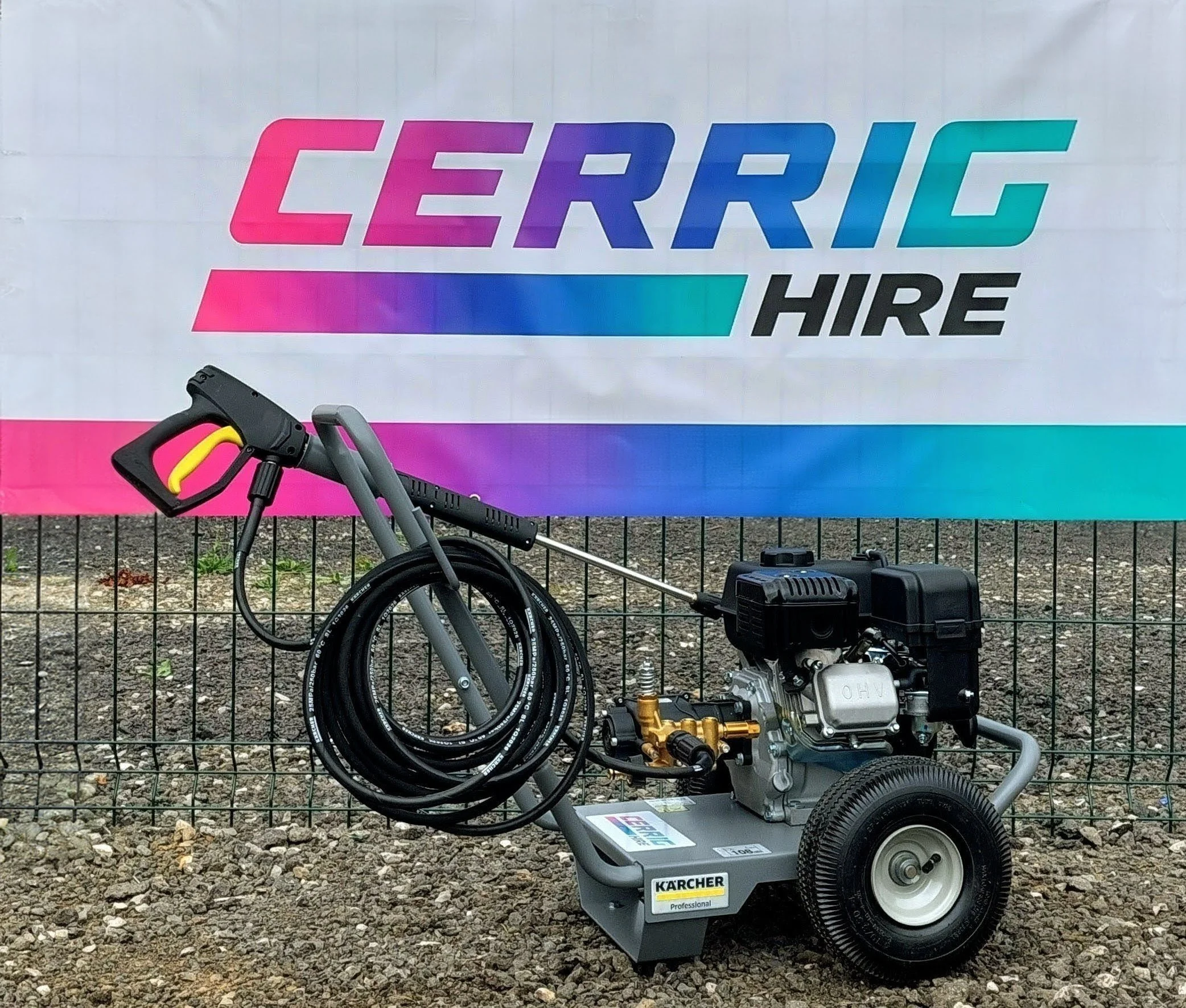 A pressure washer with a hose connected, positioned in front of a sign that reads 'CERRIG HIRE' with a gradient color background from pink to teal. The sign is mounted on a fence, and the ground is gravel.
