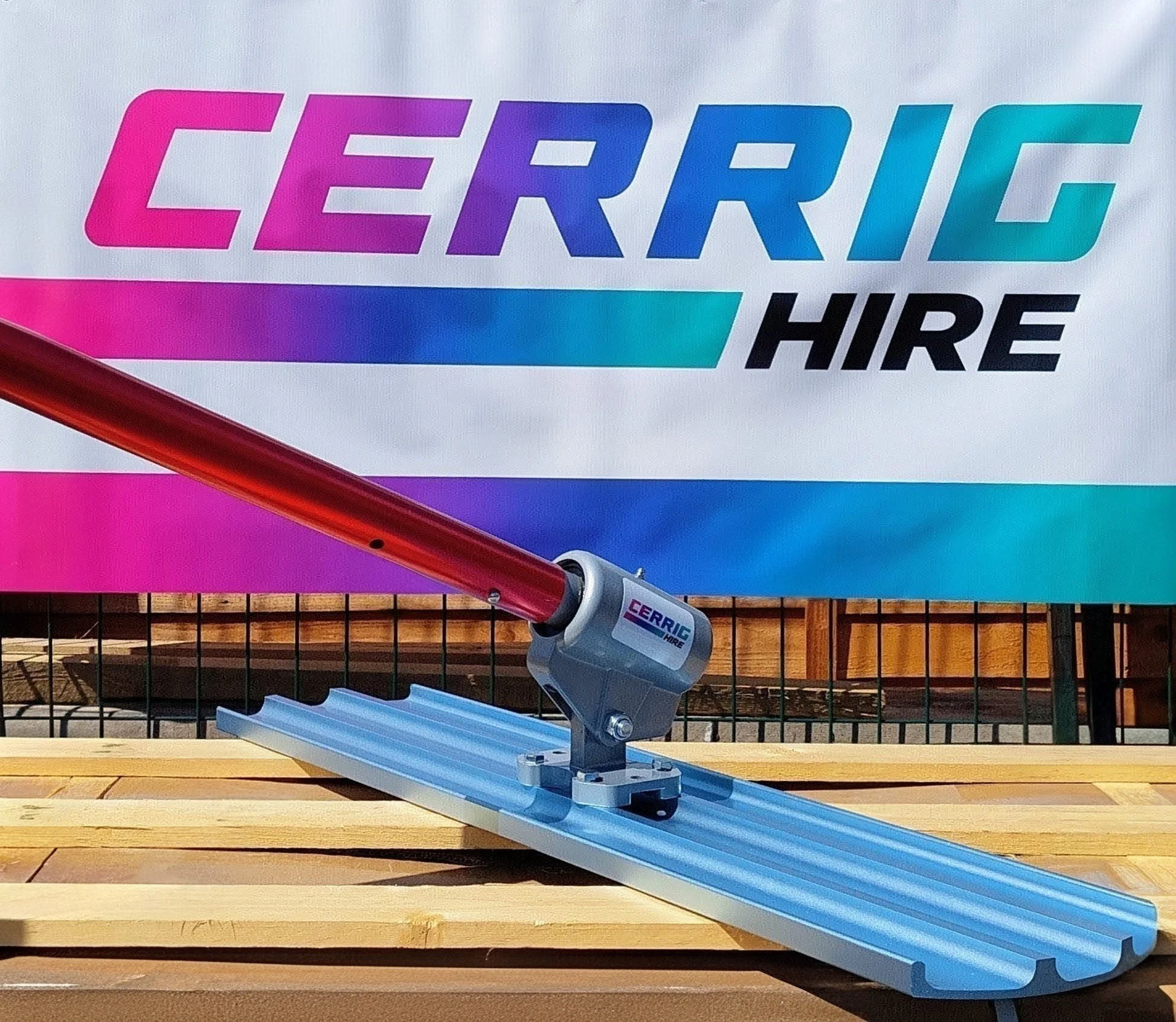 A metal bull float, resting on a rectangular wooden surface. In the background, there is a colorful sign that reads 'CERRIG HIRE' with purple, blue, and black text, and part of a red object and a wooden grid are visible.