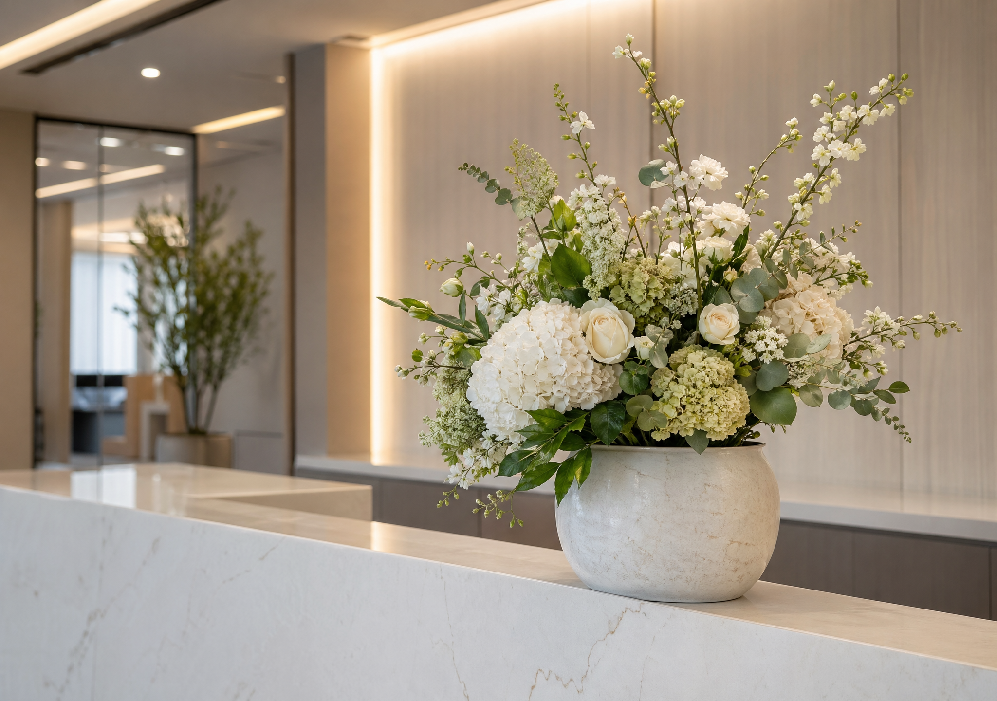 A white ceramic vase filled with white roses, hydrangeas, and other white and green flowers on a marble countertop in a modern interior space.
