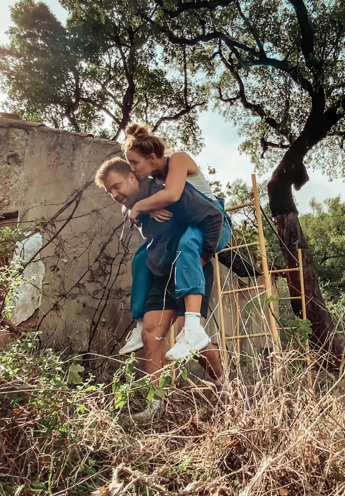 A young woman giving a piggyback ride to a man outdoors, with trees and a ladder structure in the background.