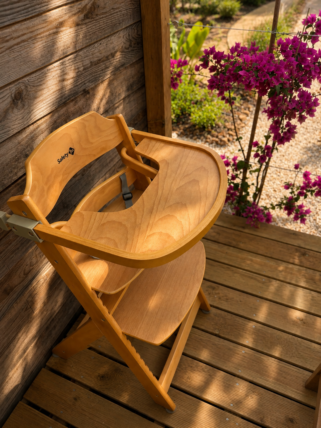 A wooden high chair on a wooden deck next to a fence, with vibrant purple bougainvillea flowers and garden plants in the background.