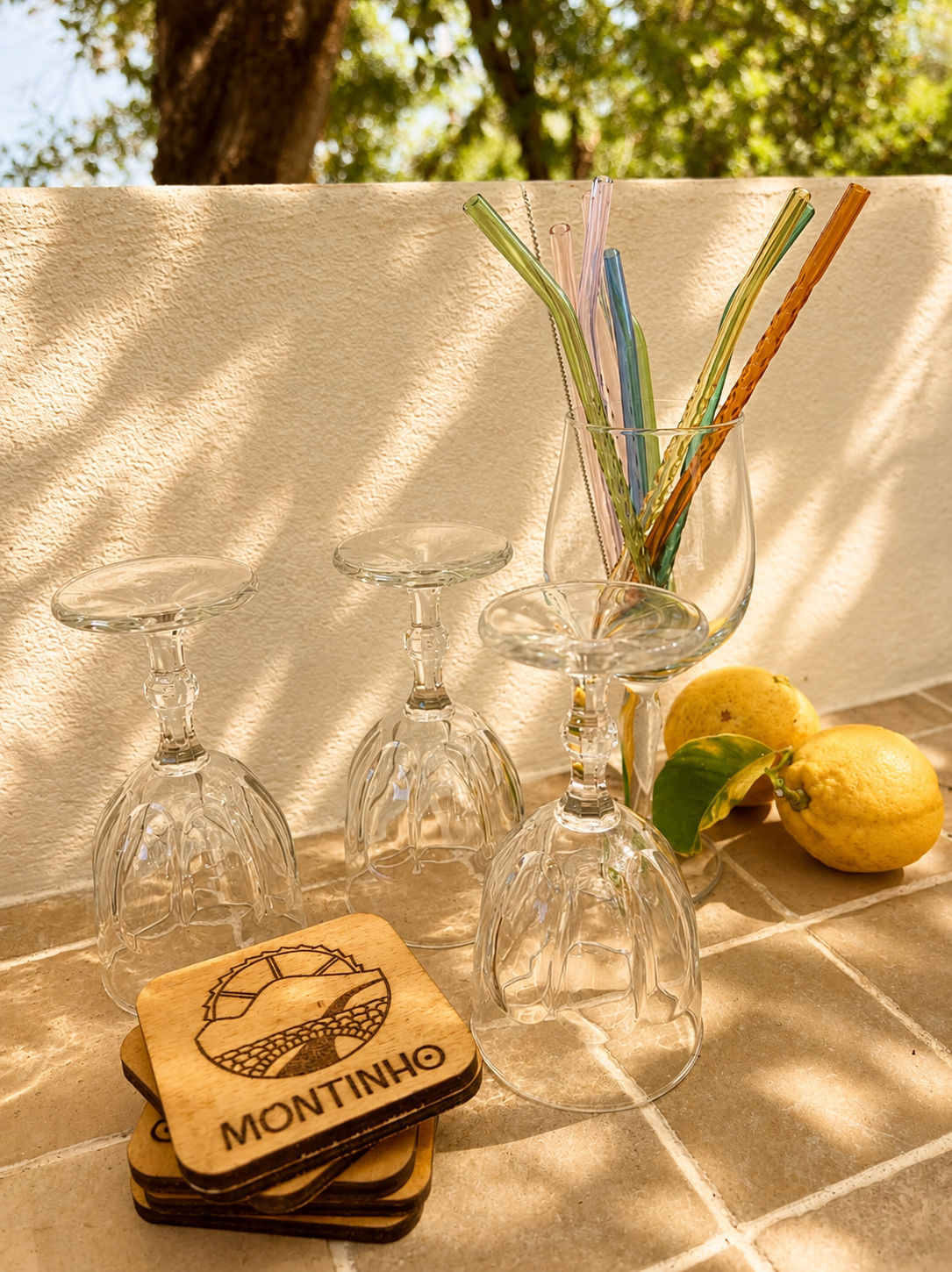 Three upside-down wine glasses, a glass with colorful straws, two lemons, green leaves, wooden coasters with a logo and text, and a patio wall with sunlight and shadows.