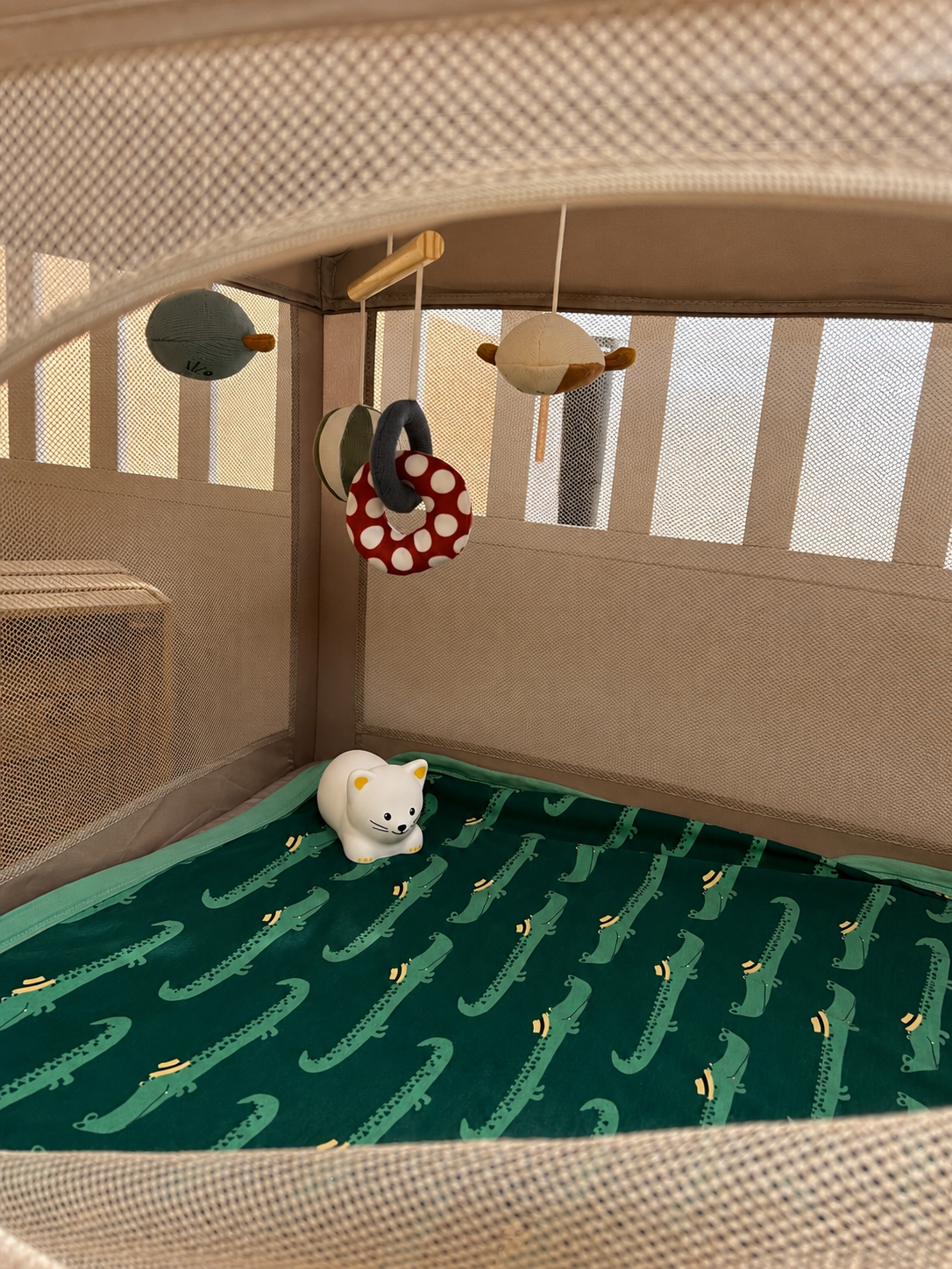 A beige playpen with a green crocodile-patterned mattress, a white cat-shaped toy, and hanging soft toys including a bird, a ring with red and white polka dots, and two other plush toys.