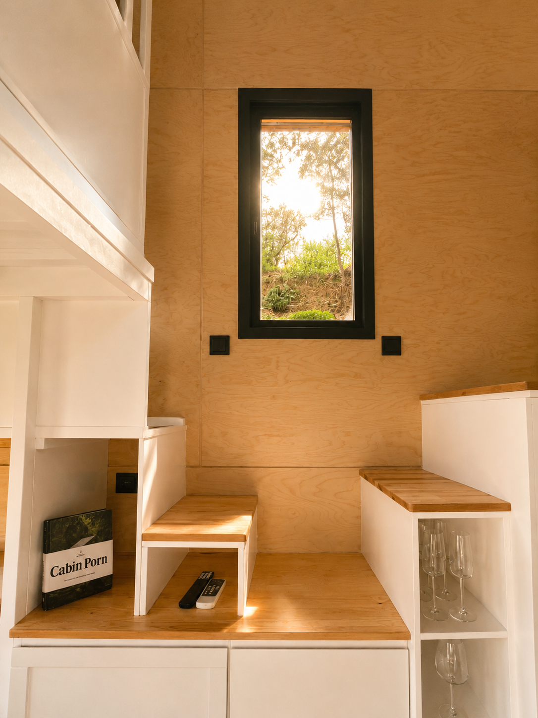 Small room with a black-framed window, wood-paneled walls, white furniture with wooden surfaces, a book titled 'Cabin Porn', a remote control, and some wine glasses on a shelf.