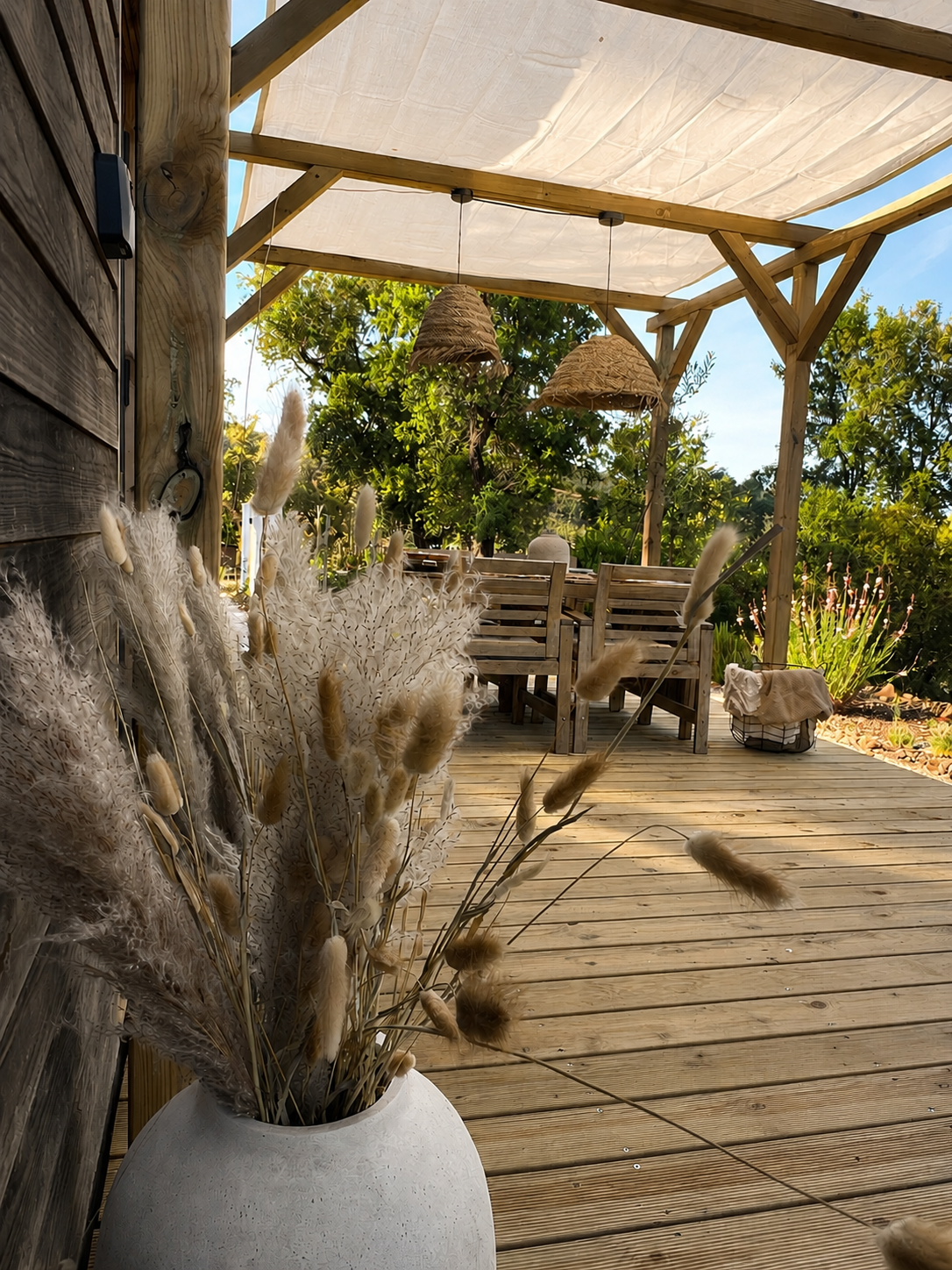 Outdoor wooden deck with dried floral arrangement in a white vase in the foreground, wooden furniture in the background, and lush greenery beyond under a partially covered shed.