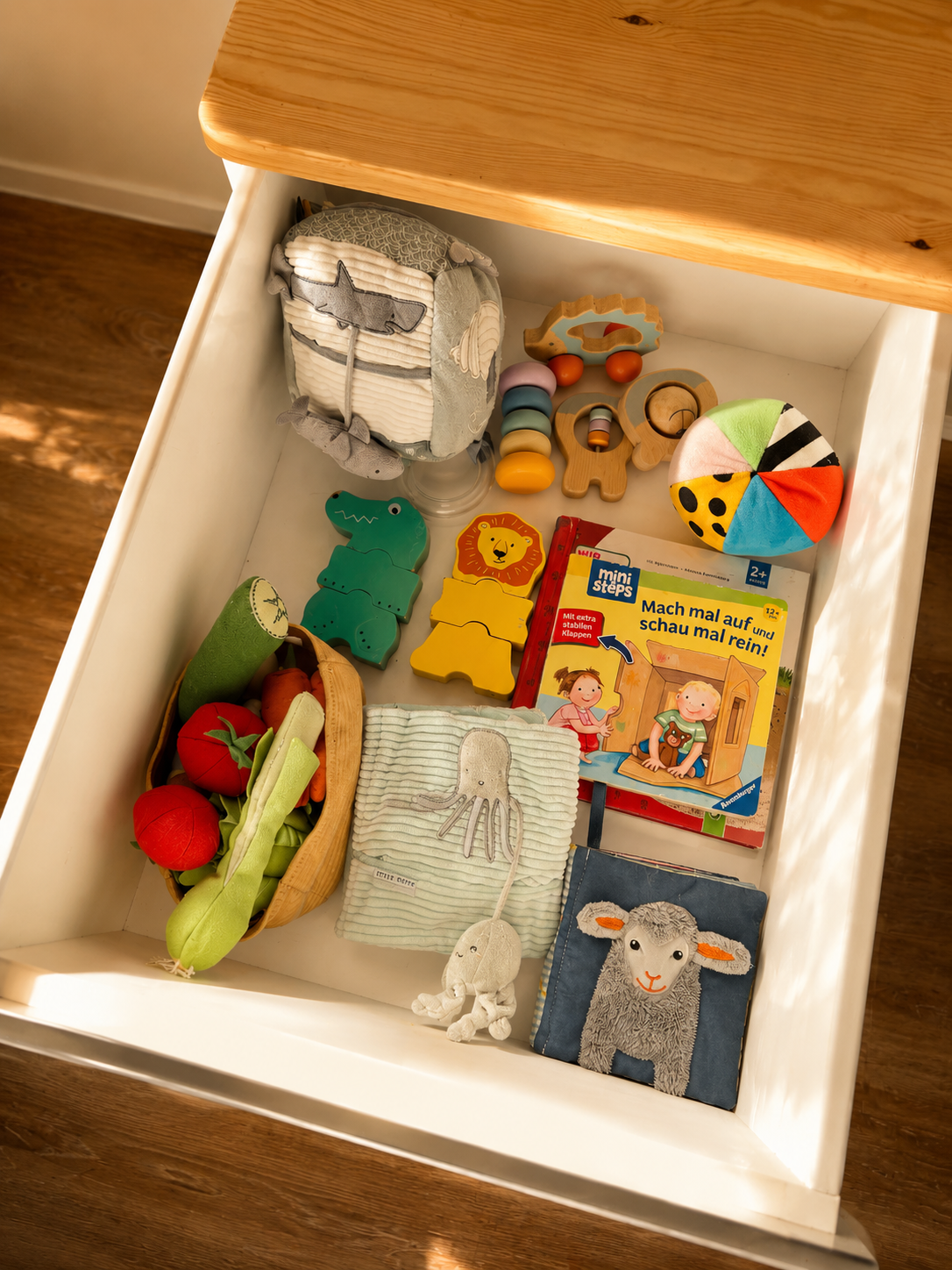 Open drawer containing children's toys, books, and cloth items, including plush vegetables, a fabric toy with a tentacle design, animal-shaped stacking toys, a colorful fabric ball, a wooden puzzle, a children's book in German, and fabric plush animals.