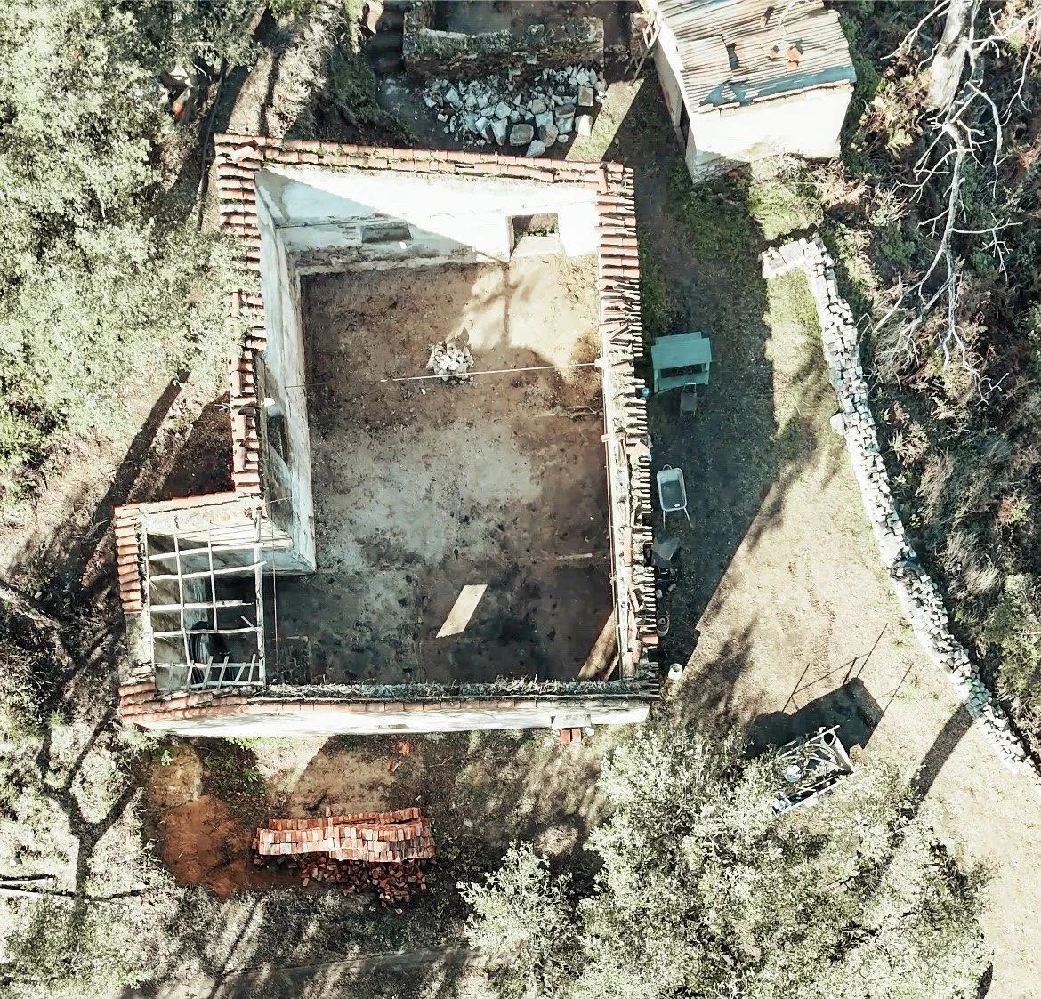 An aerial view of a house under construction with walls partially built, construction materials, and equipment in a backyard surrounded by trees and a stone border.
