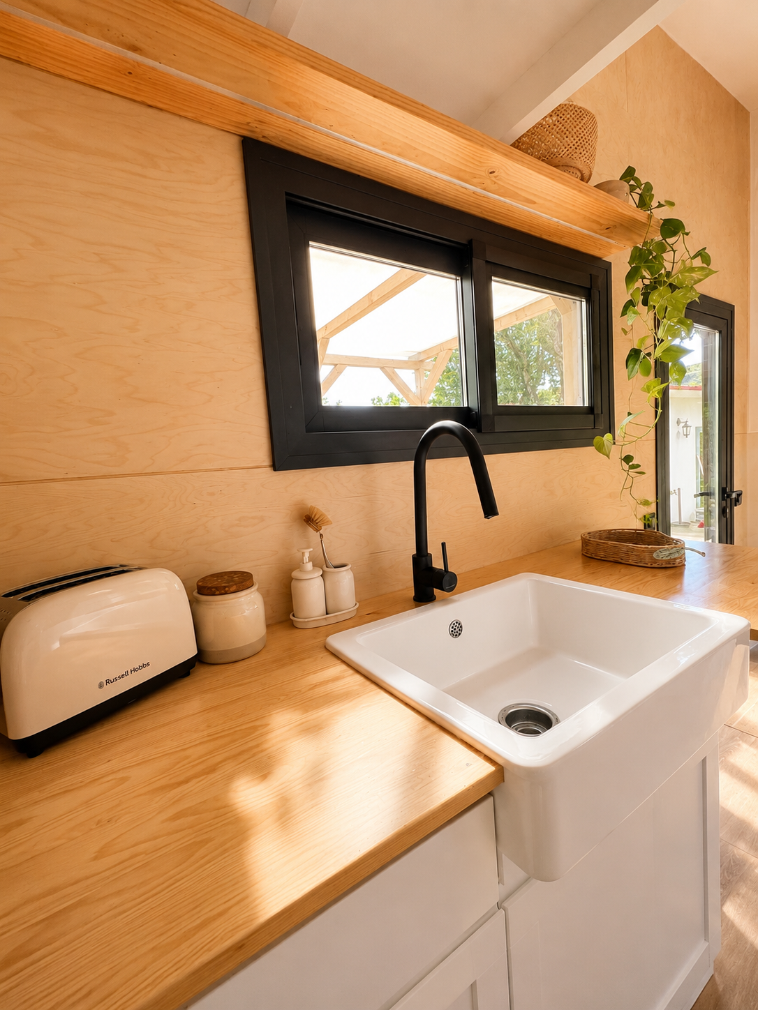 A wooden countertop kitchen sink area with a black faucet, a window with black trim, a Russell Hobbs toaster, ceramic jars, soap dispensers, and a plant trailing from a shelf.