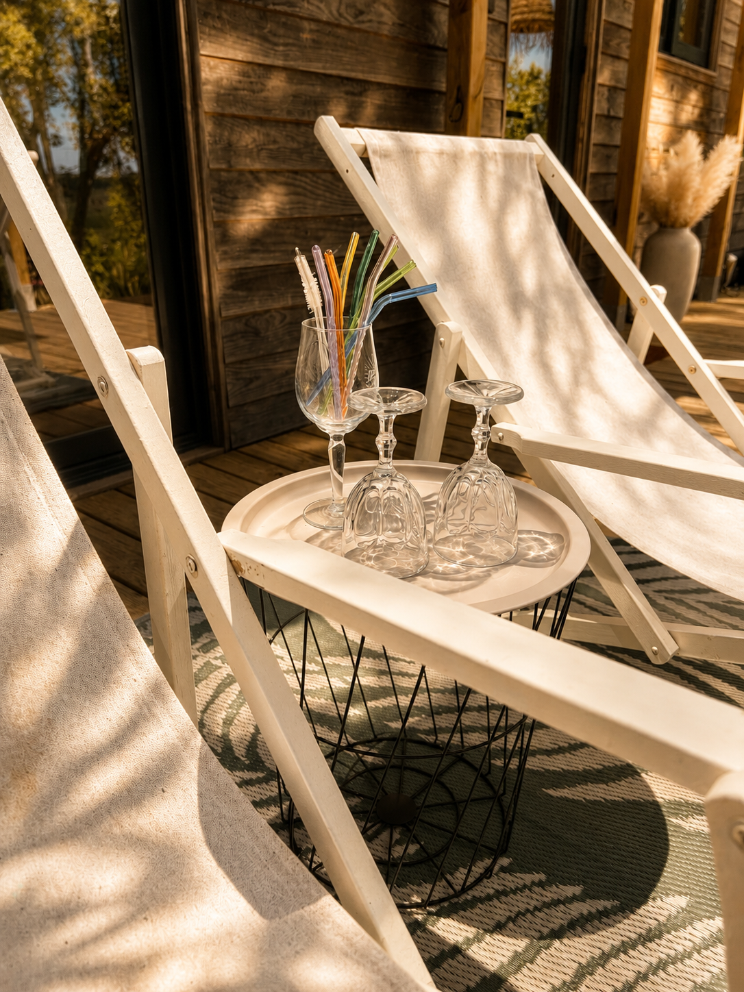 Outdoor patio scene with two white deck chairs, a round wire side table, and a tray holding two upside-down wine glasses, a champagne flute, and a vase with drink stirrers, on a patterned outdoor rug, with a wooden wall and windows in the background.