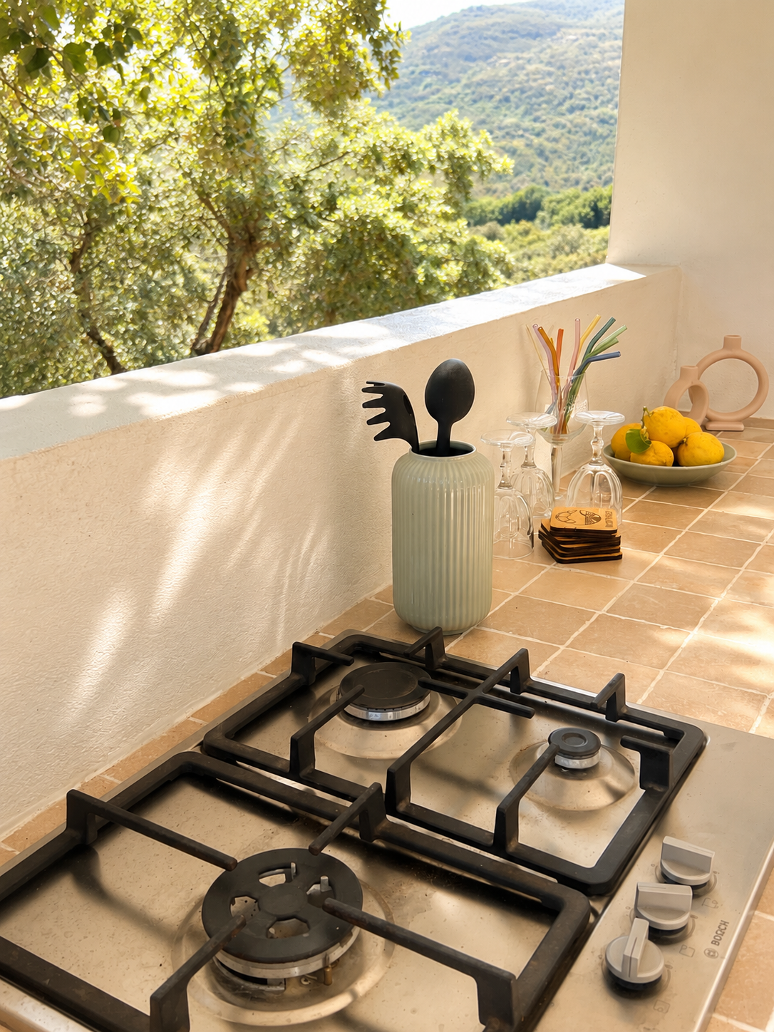 Kitchen counter with a gas stove, a ceramic utensil holder with black utensils, upside-down glasses, a bowl of lemons, colorful straws, and a mountain view outside.