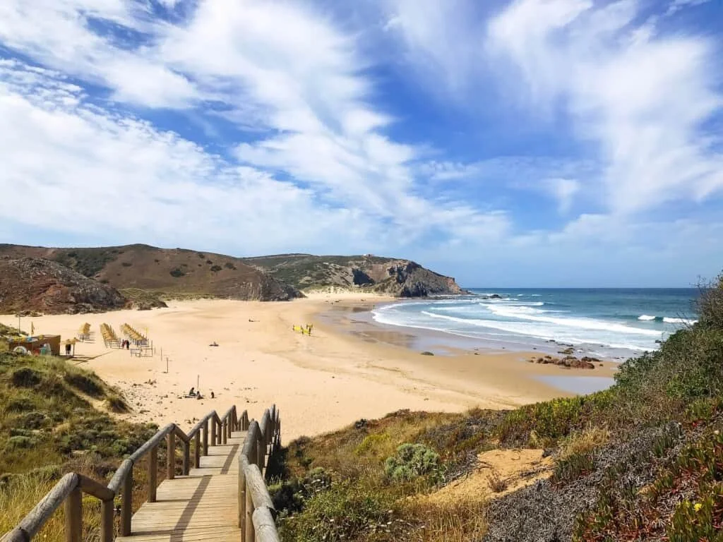 A sandy beach with a wooden staircase leading down to the sand, overlooking the ocean with waves and cliffs in the background under a blue sky with clouds.