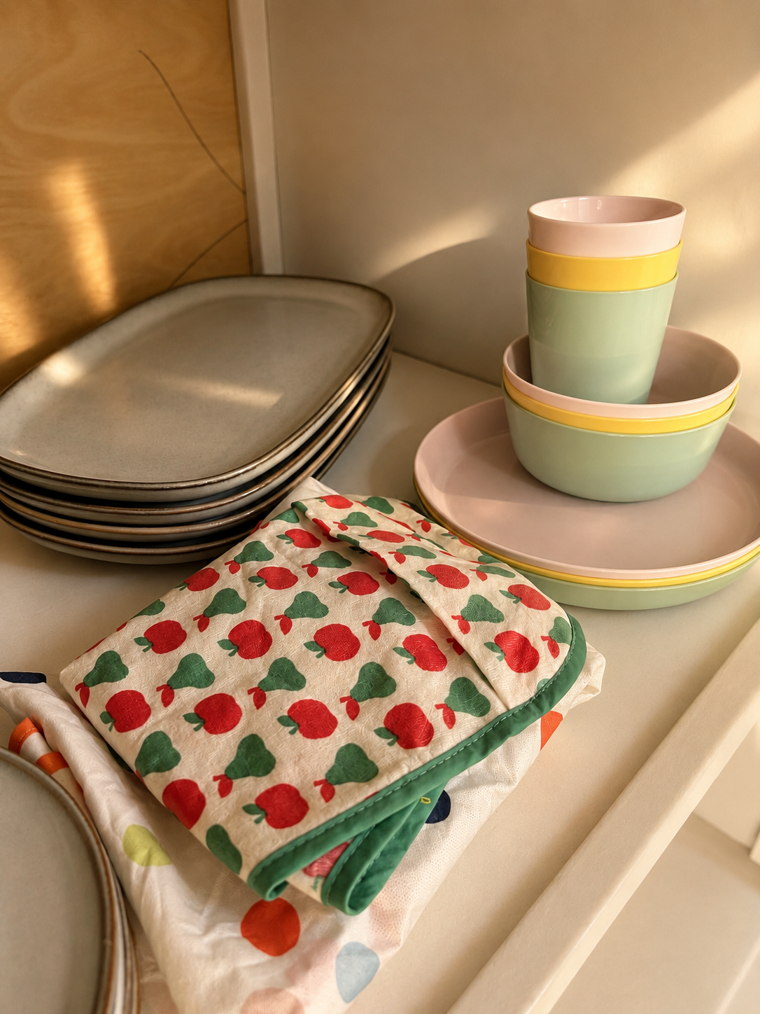 Stacked pastel bowls, black and gray oval plates, a patterned oven mitt with red and green apples, and a mix of white and patterned dishes on a kitchen counter.
