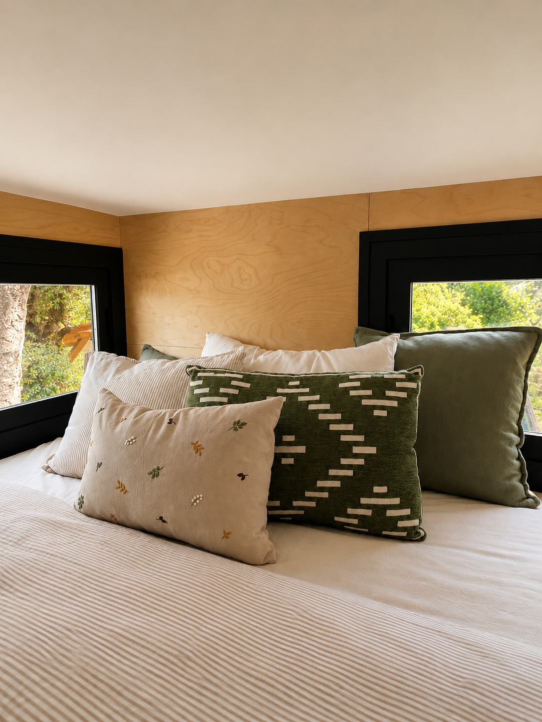 A cozy bed with multiple decorative pillows in a bedroom, featuring black-framed windows and light wooden walls, with a view of green trees outside.