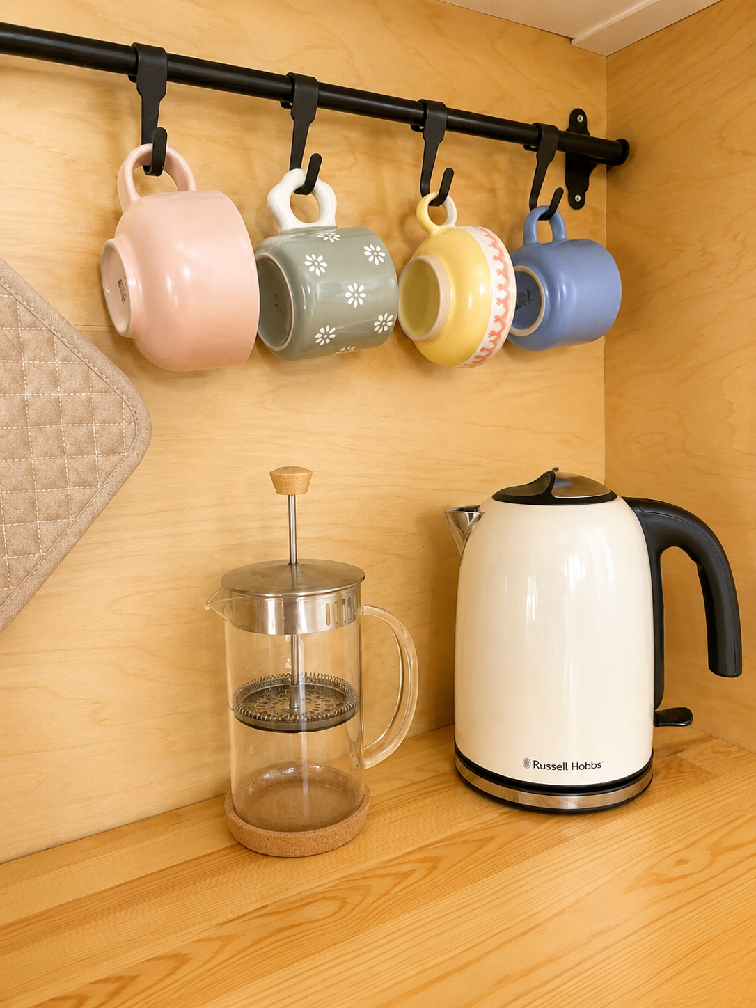 Four colorful coffee mugs hanging on a black rod, a French press on a wooden countertop, and an electric kettle with a cream body and black handle on a wooden wall.