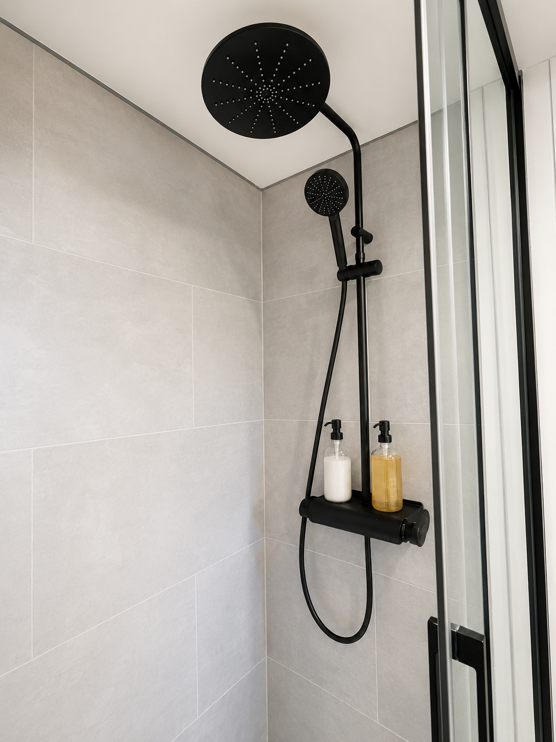 Modern shower with black rainfall showerhead, hand shower, gray tiled walls, and two soap dispensers.