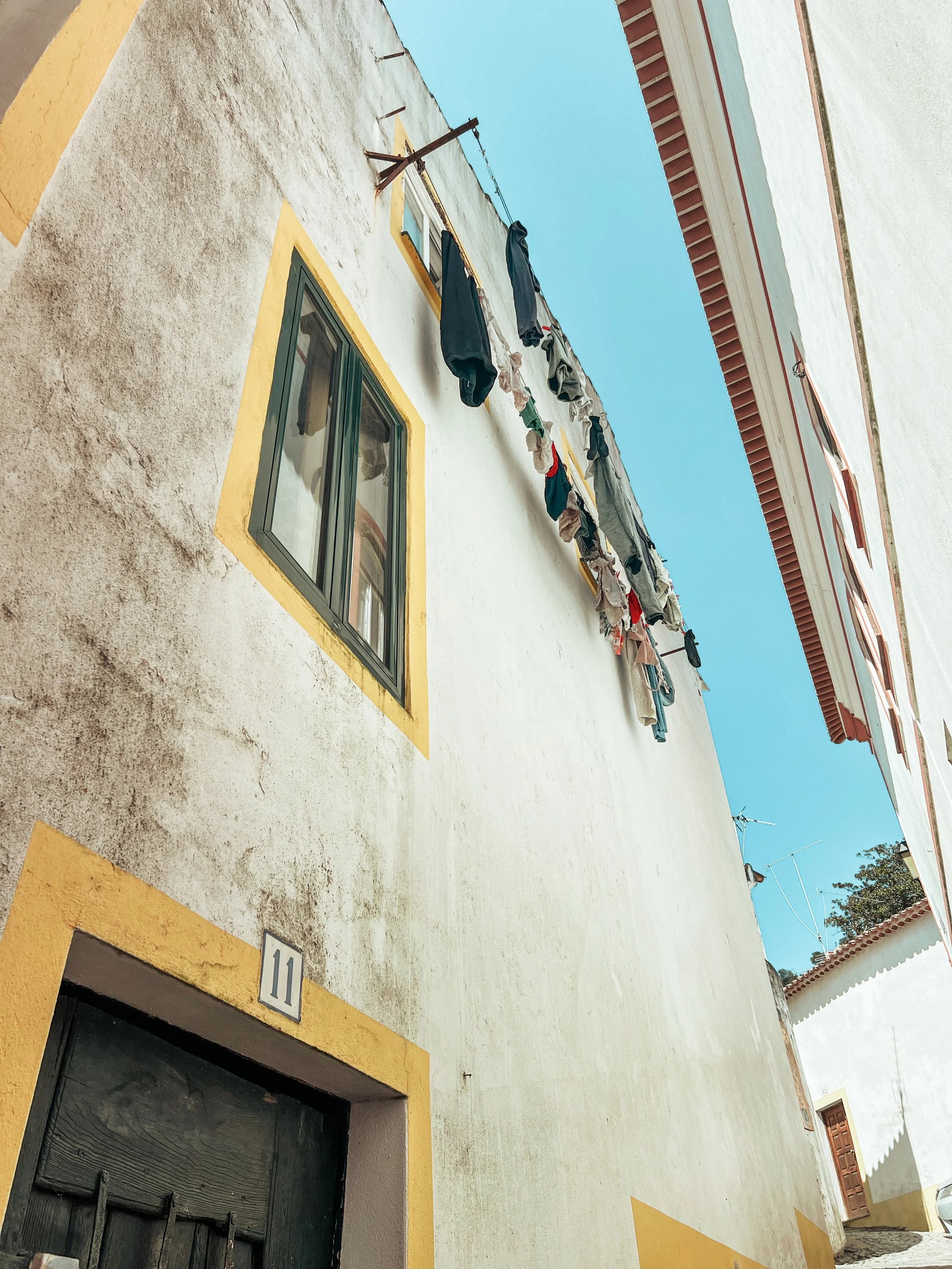 A white building with yellow accents around the window and door, with laundry hanging on a clothesline outside. The sky is blue and clear.