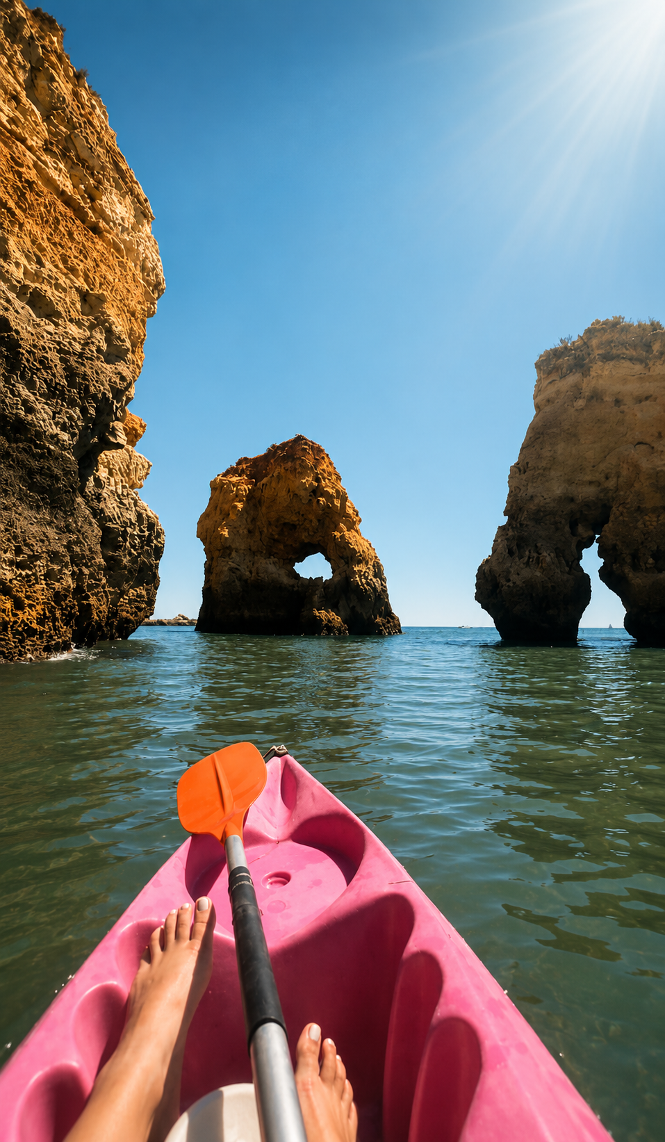 View from a pink kayak with a paddle, paddling through water surrounded by large rock formations with numerous holes, under a clear blue sky with the sun shining.