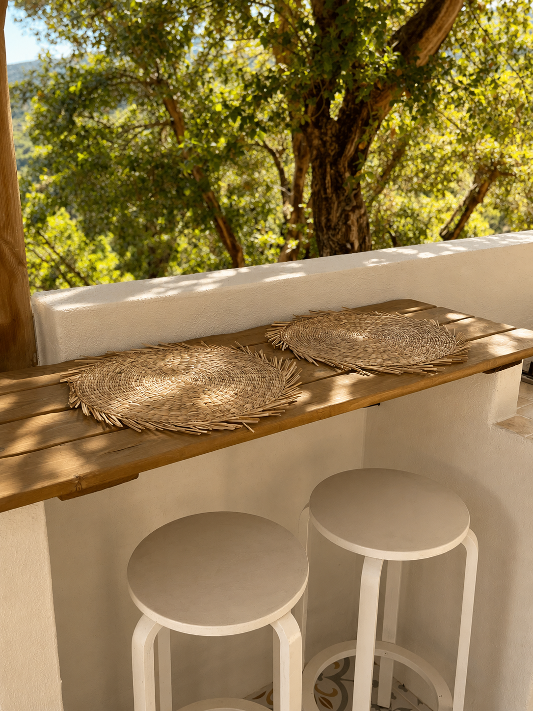 A balcony with two white barstools, a wooden counter with two woven placemats, white stucco wall, and a view of a leafy green tree outside.