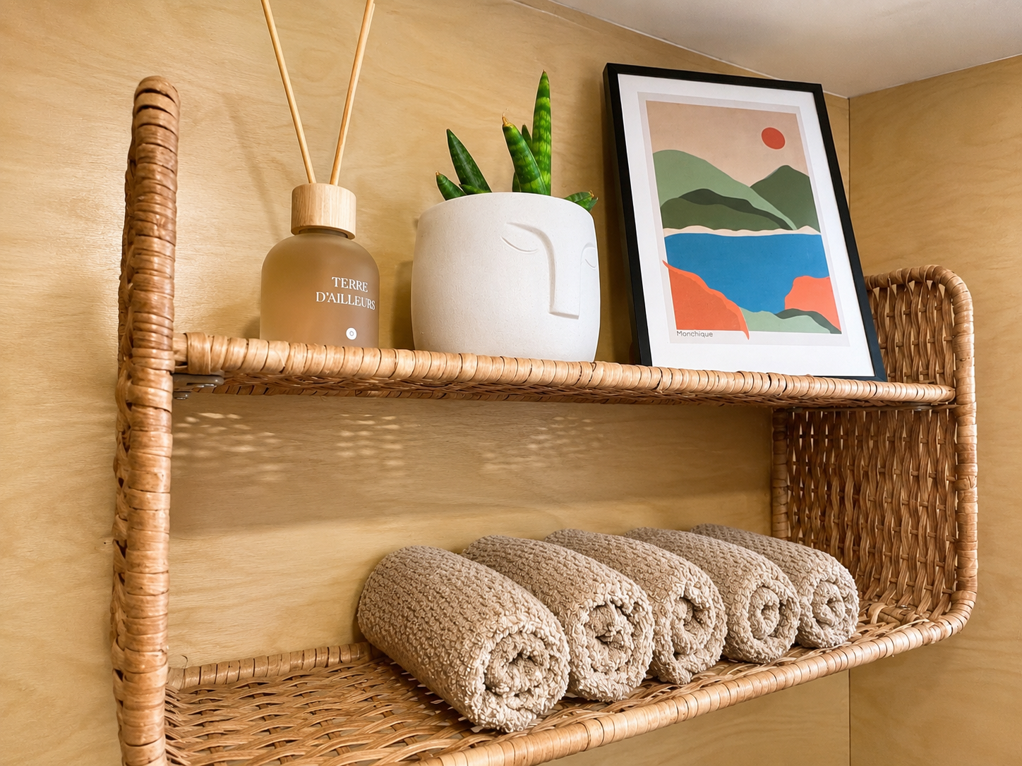 A wicker wall shelf holding rolled beige towels, a white vase with a face design and green plants, a framed landscape art print, and a bottle with reed diffuser sticks, all against a light wood wall.