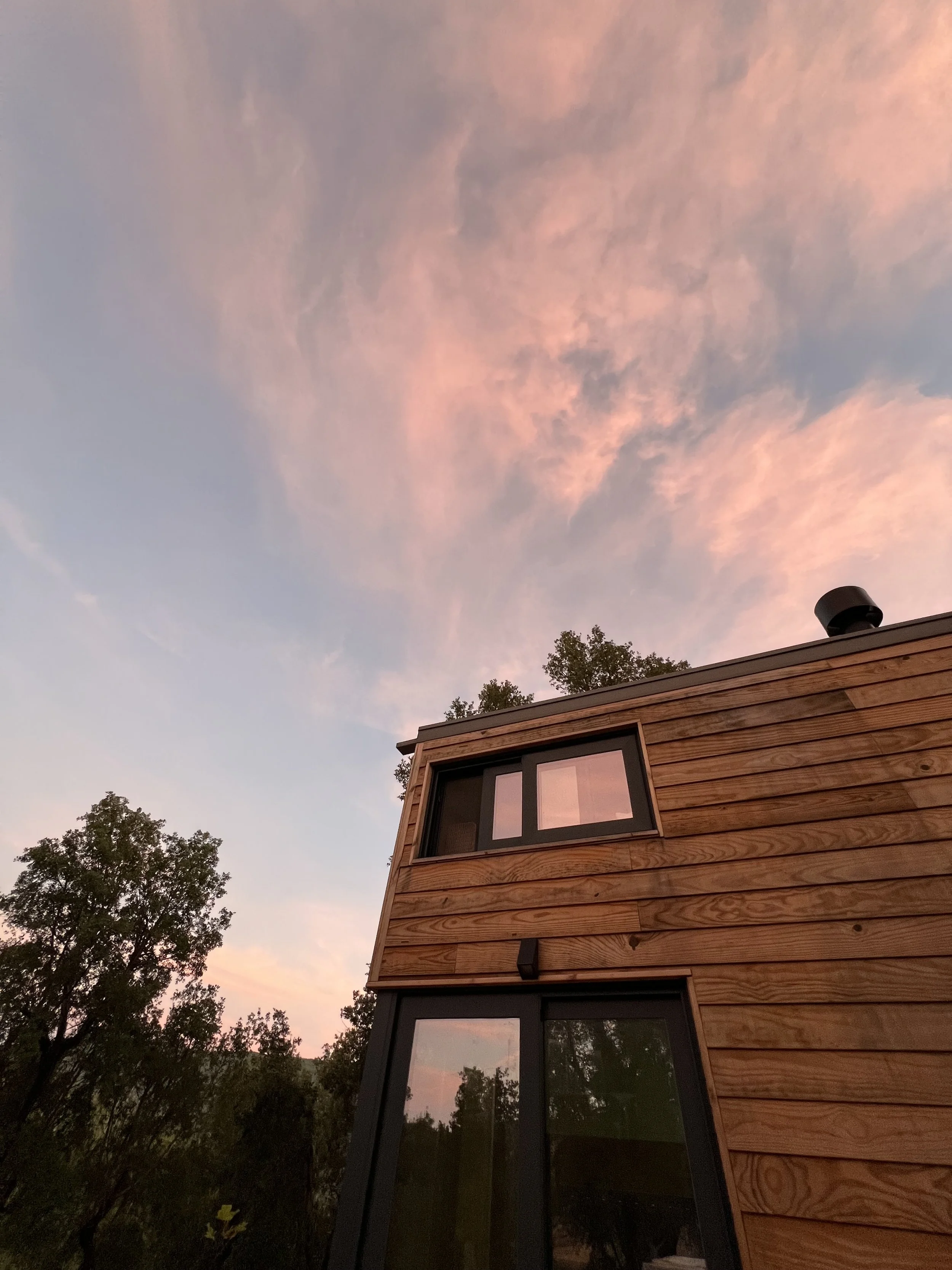 A modern wooden house with large windows reflecting a pink and purple sky at sunset, surrounded by trees.