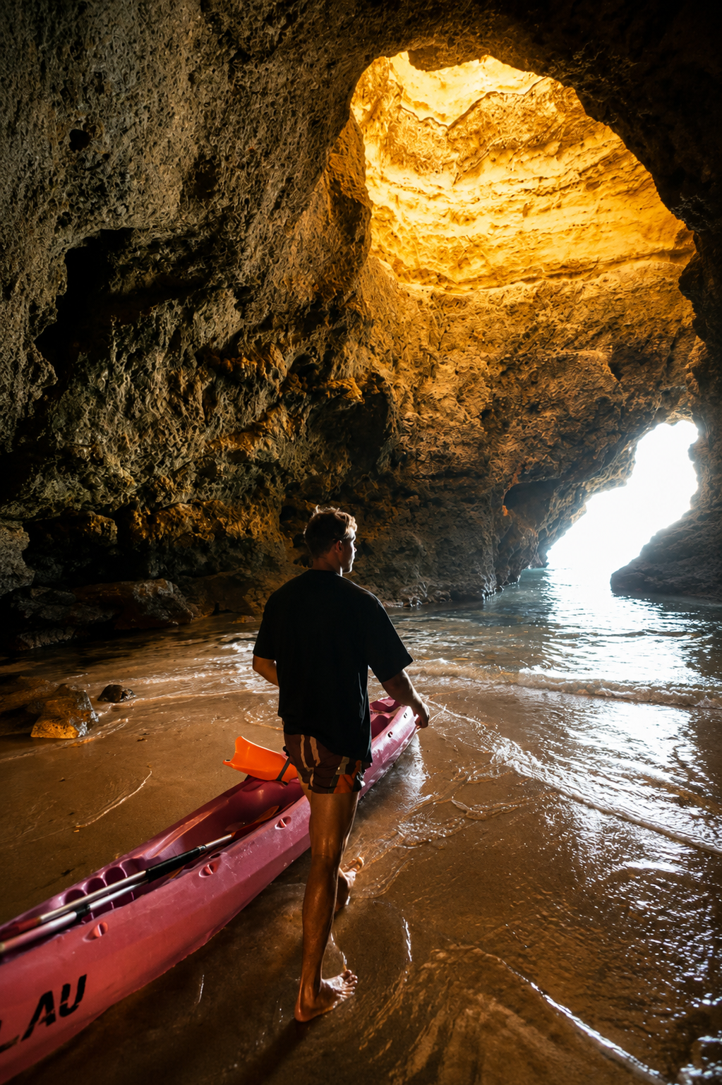 A person carrying a kayak on a sandy beach inside a dark rocky cave with sunlight shining through an opening at the end of the cave
