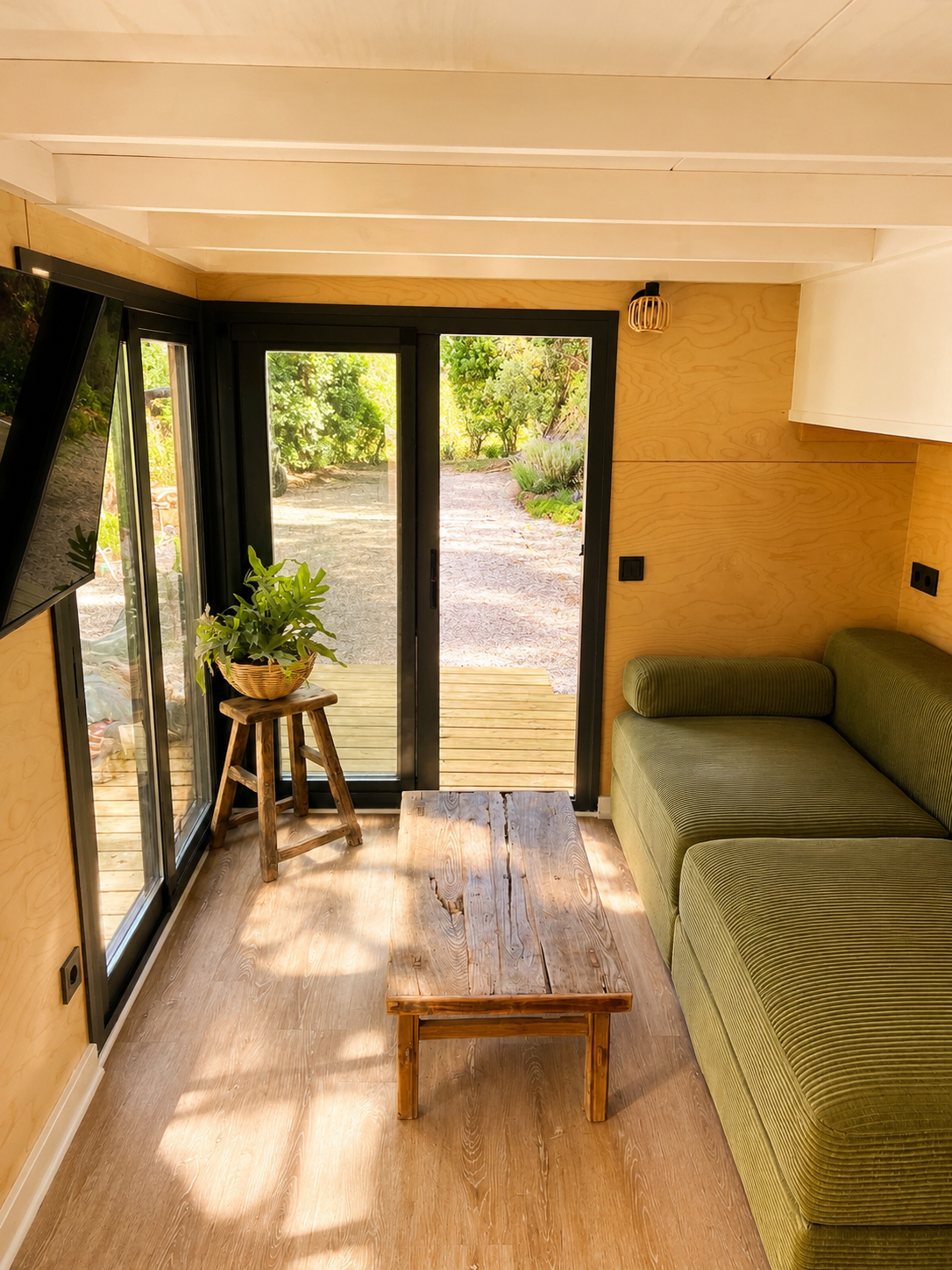 Interior of a cozy room with wooden walls and ceiling, a sliding glass door leading outside to a wooden deck, a green sofa, a rustic wooden coffee table, a small wooden stool with a potted plant, and a flat-screen TV mounted on the wall.