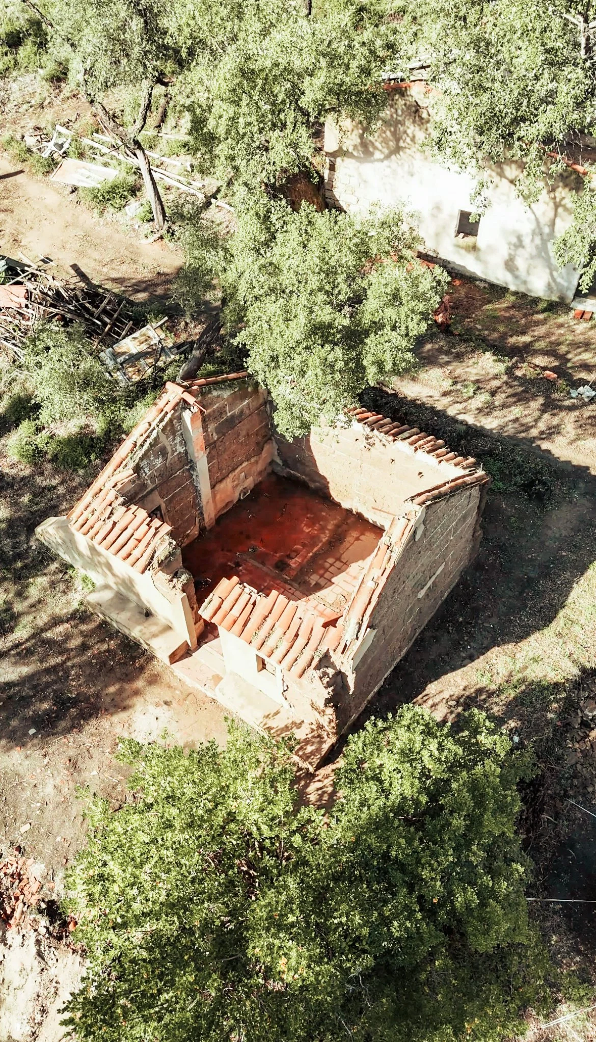 An aerial view of a partially destroyed house with exposed brick walls and a red dirt floor, surrounded by green trees and other debris.
