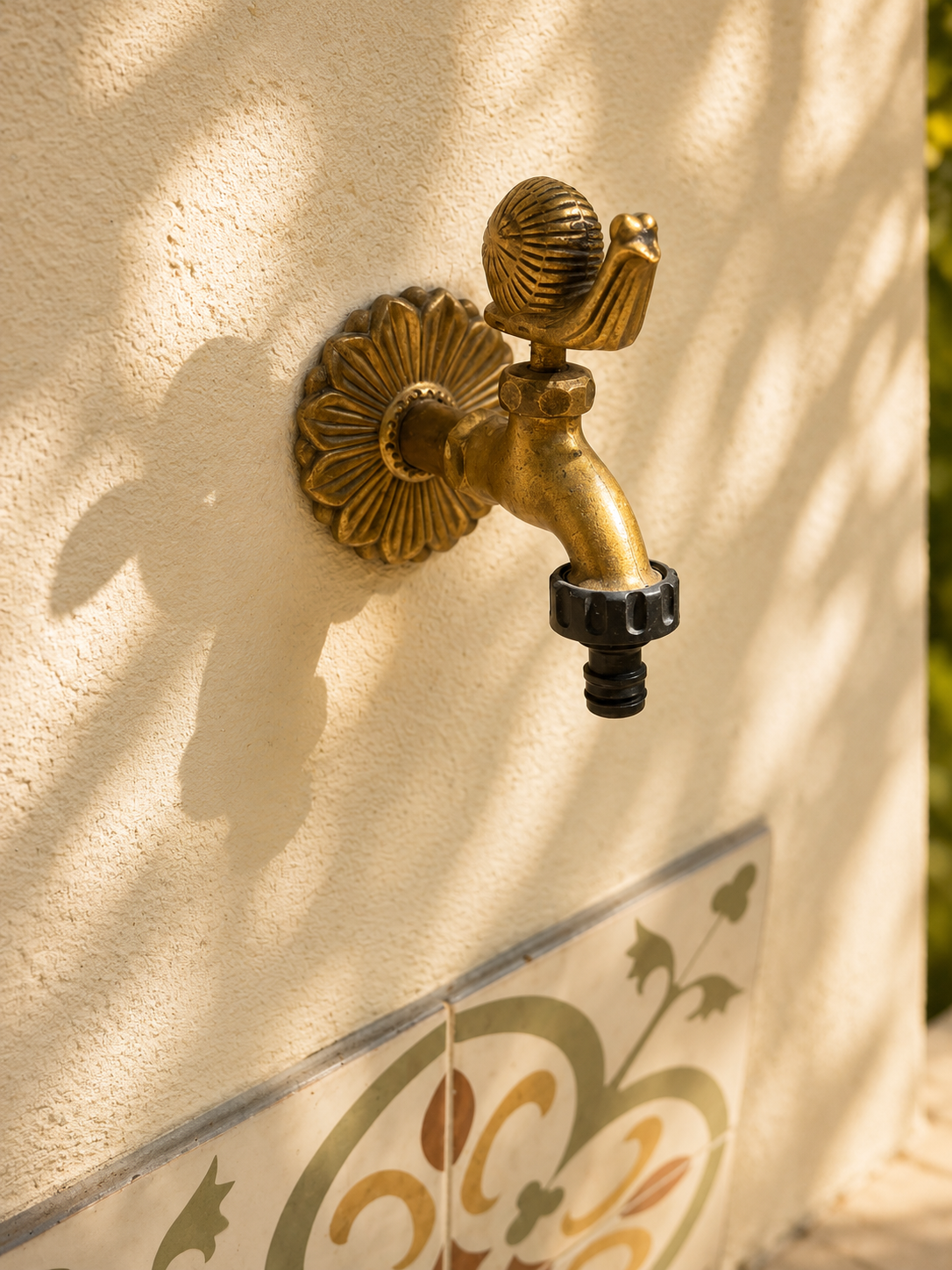 Close-up of a brass outdoor water faucet attached to a textured beige wall, with decorative ceramic tiles below and soft shadows cast on the wall.