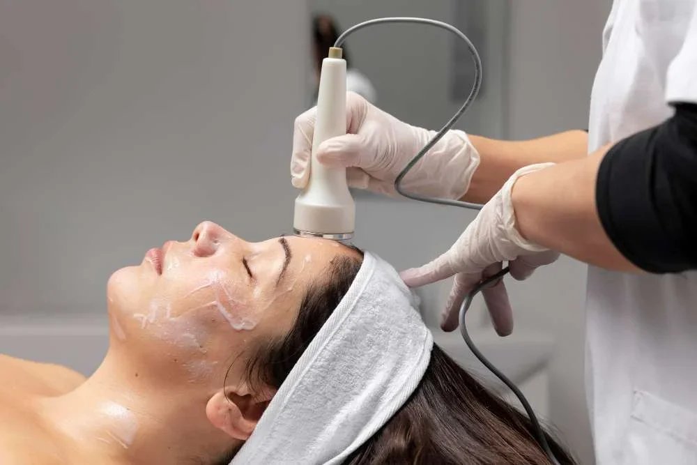 A woman lying down with her eyes closed is receiving a facial treatment with an ultrasound device from a professional wearing gloves.