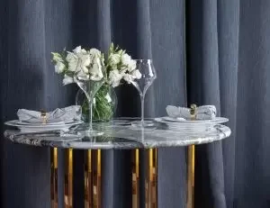 Round dining table with a floral centerpiece, set with plates, silverware, and drinking glasses, against a dark curtain background.