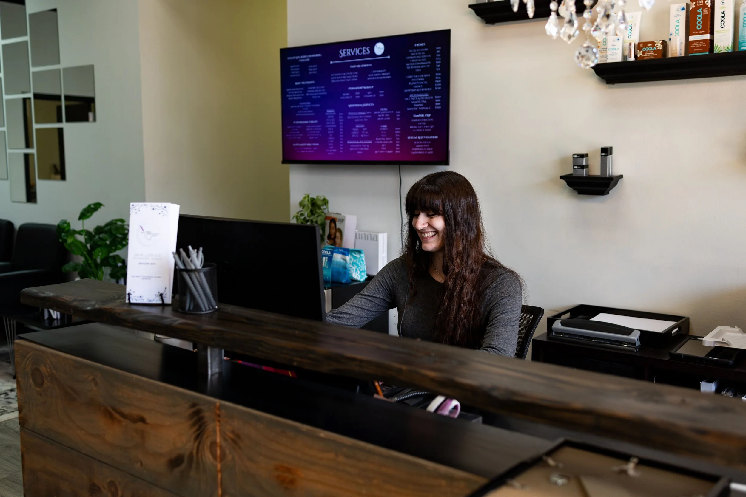 A woman with long dark hair smiling at a computer behind a wooden reception desk in a modern lobby at Arctic Medical Spa
