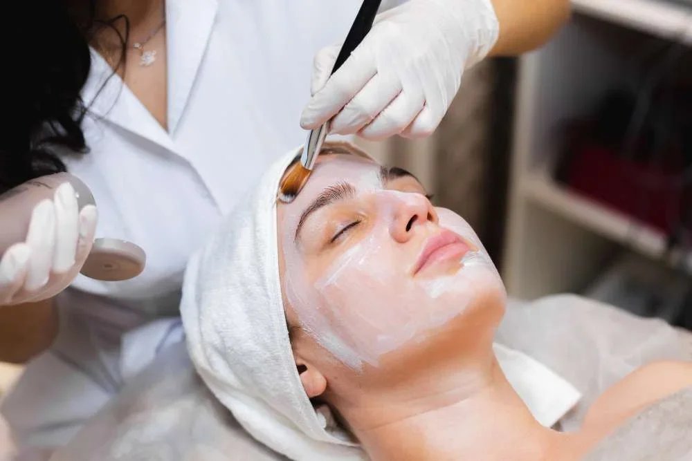 Person receiving a facial treatment at a spa or skincare clinic with the skincare professional applying a face mask or treatment.